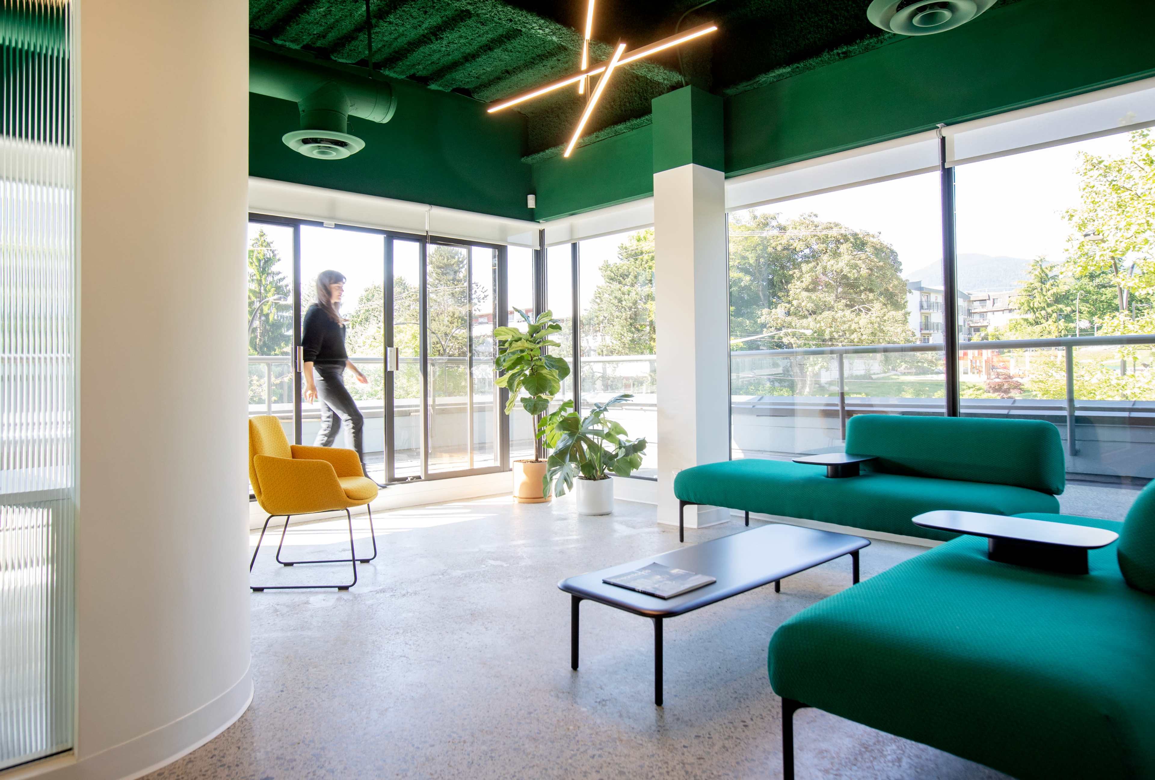 A person walks through a modern lounge area featuring green walls, large windows, and contemporary furniture including a yellow chair and teal sofas.