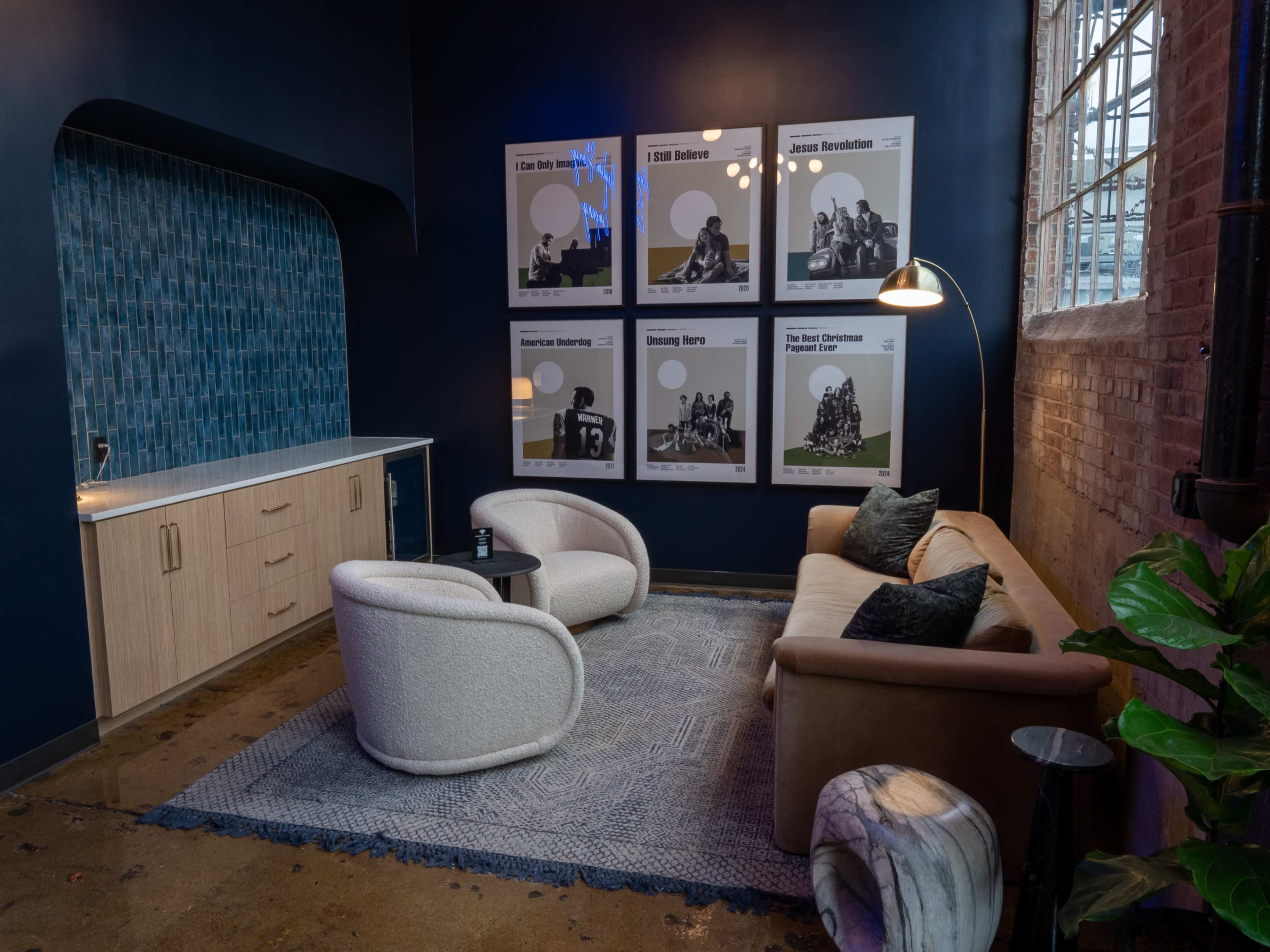 The image shows a modern lounge area with two curved chairs, a beige sofa, a round coffee table, a tall lamp, and framed black-and-white posters on the wall.
