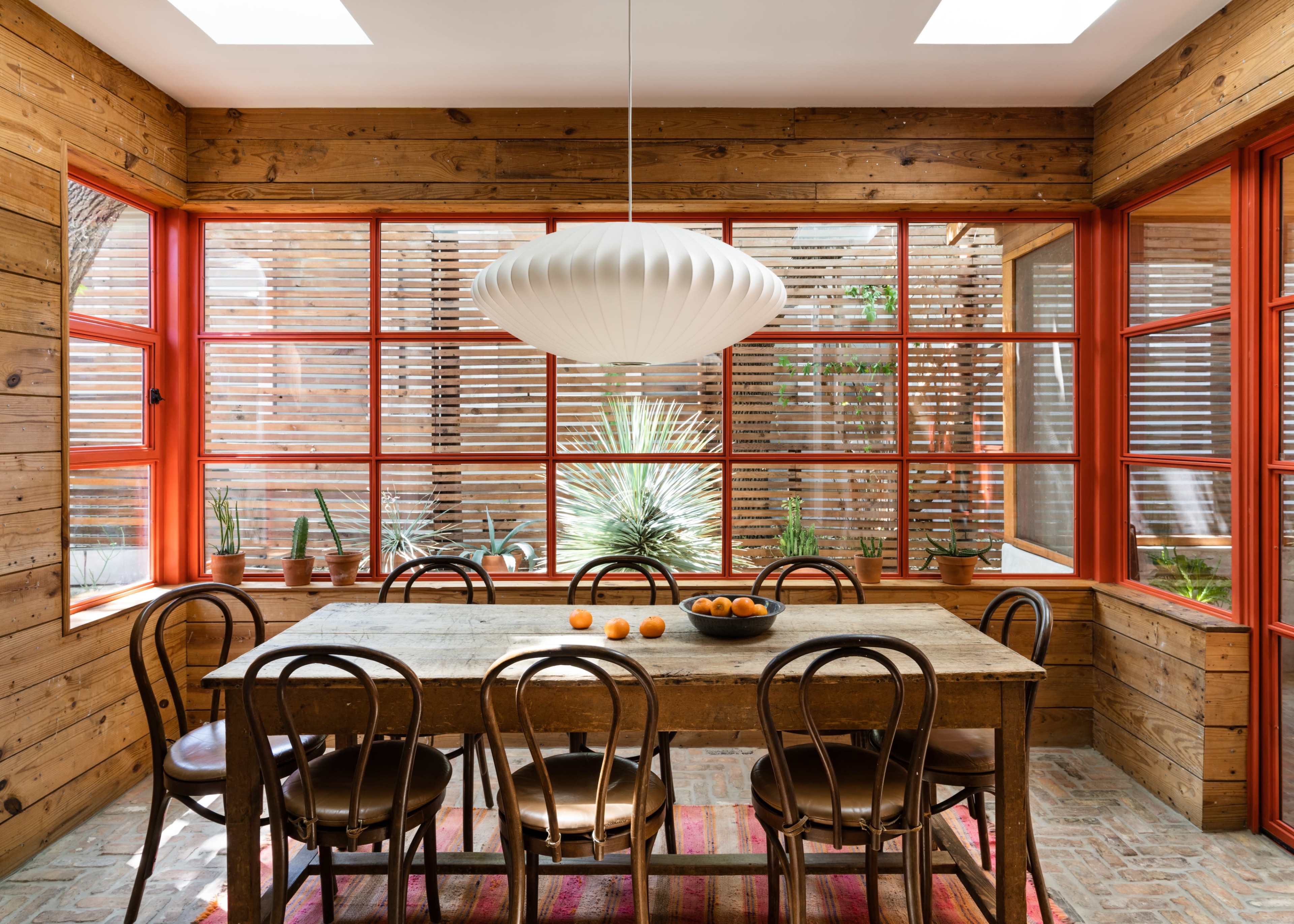 A wooden dining table surrounded by chairs is situated in a bright room with large windows and orange trim, featuring a bowl of oranges in the center.