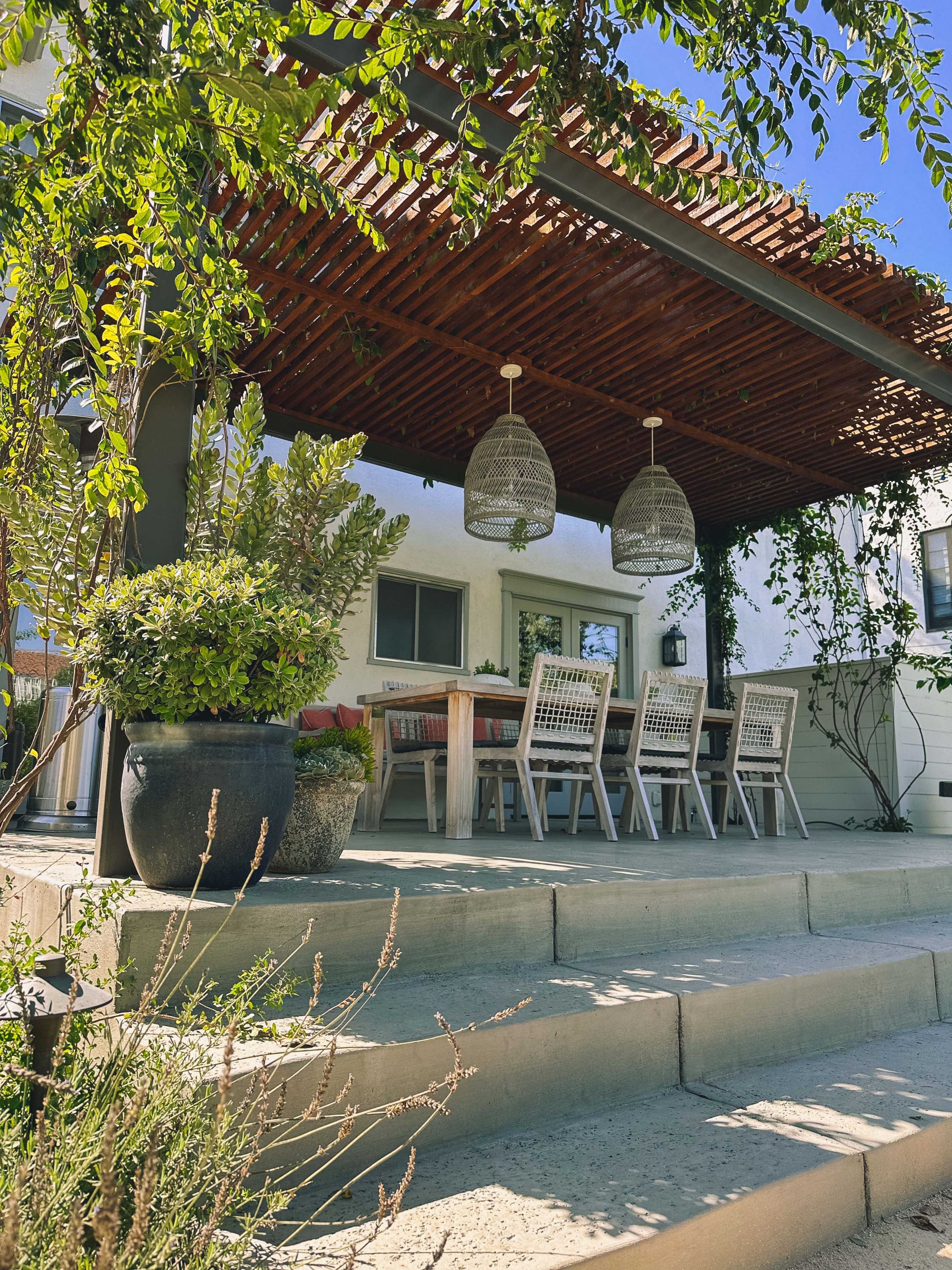 A covered outdoor patio features a dining table and chairs, surrounded by potted plants and steps leading up to a house.