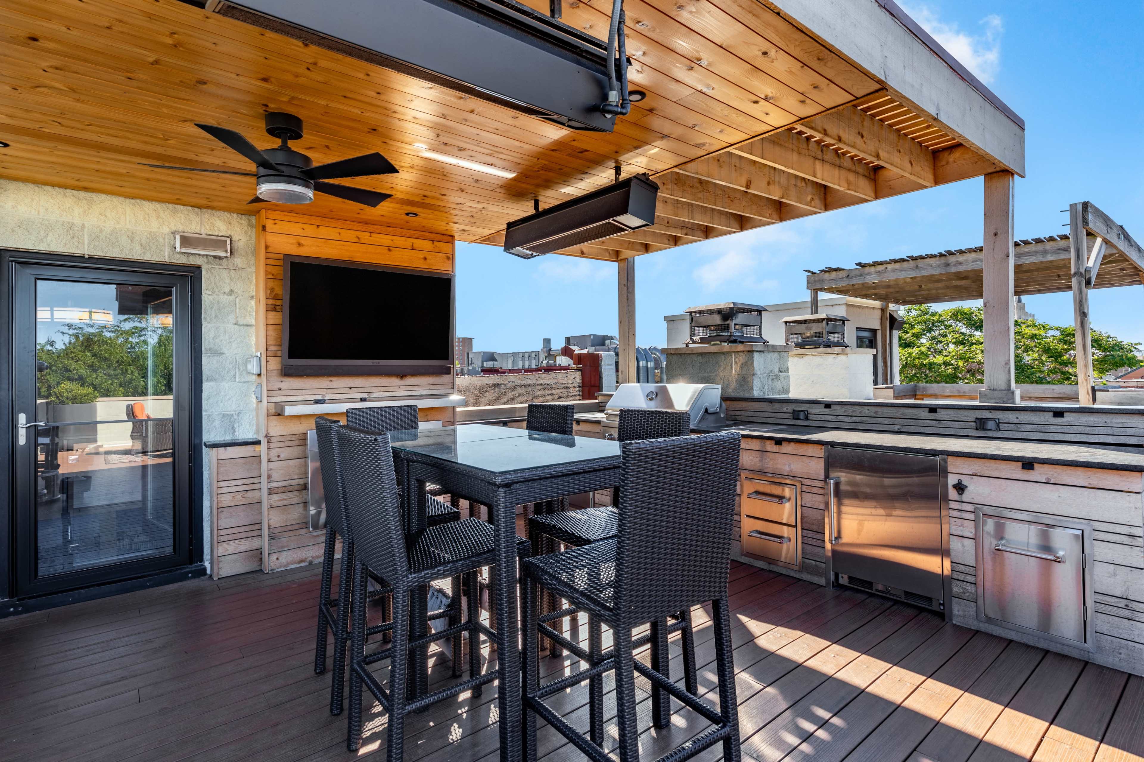 The image shows a spacious outdoor rooftop terrace featuring a dining table, high chairs, a grill, and a television under a wooden canopy.