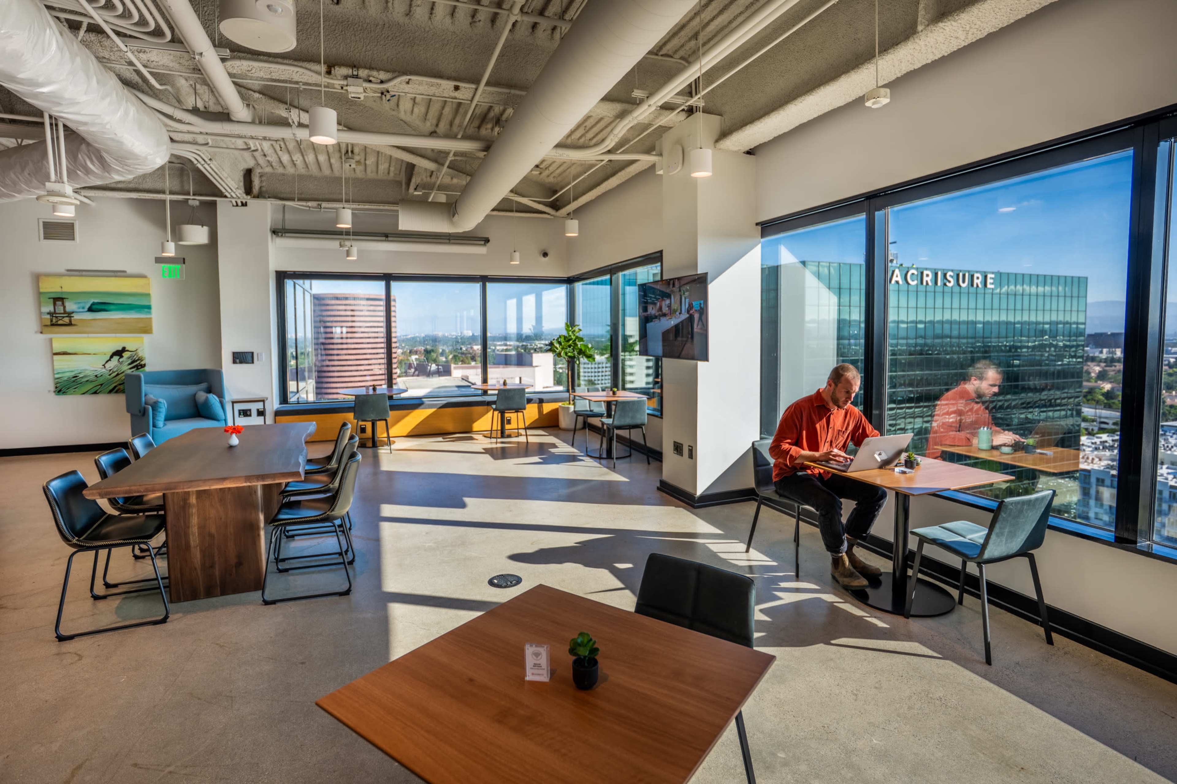 A man sits at a table using a laptop in a modern office with large windows offering a view of a cityscape.