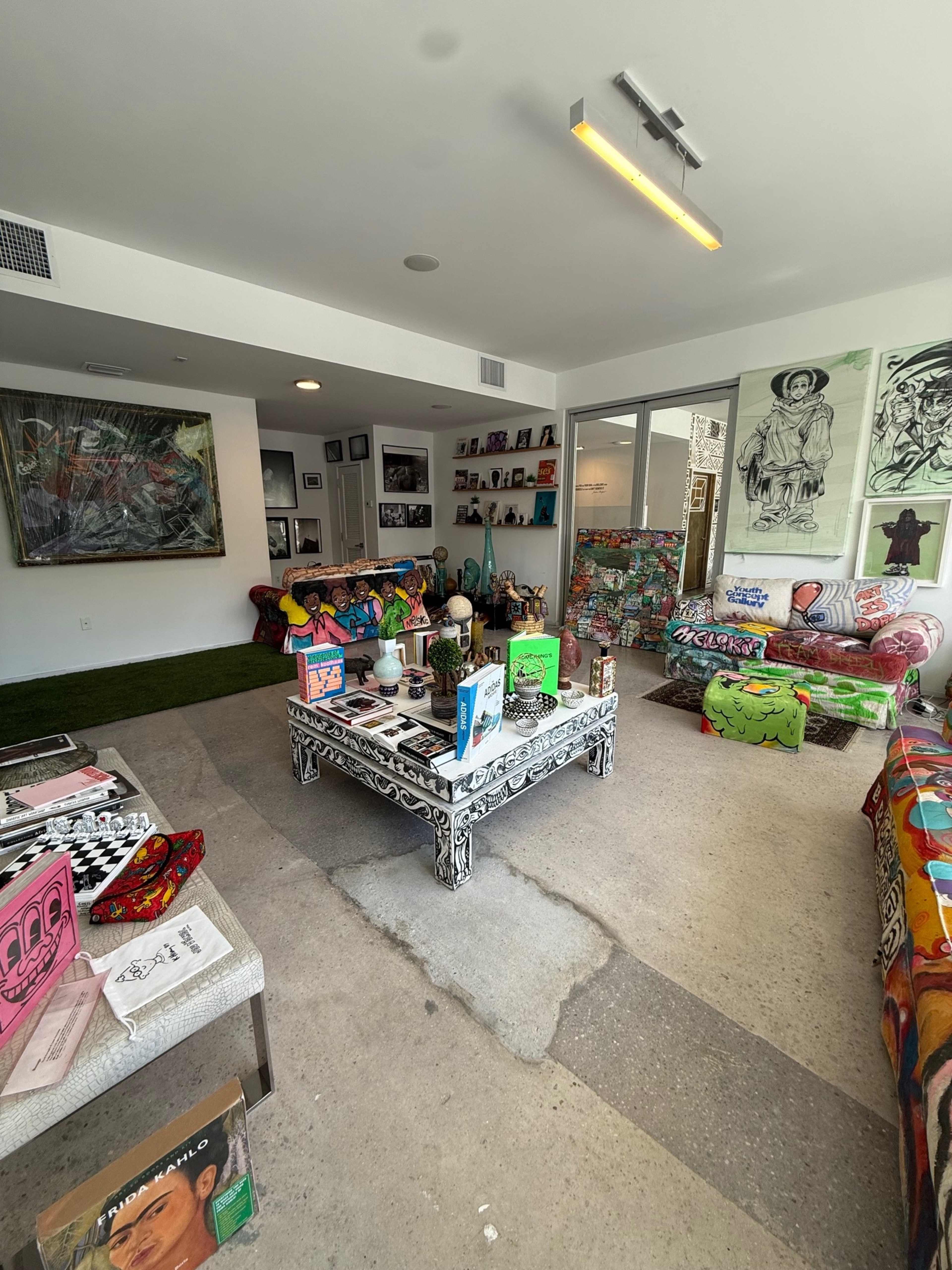 The room features colorful furniture and artwork, with a coffee table cluttered with books and objects, and a grass carpet in the center.