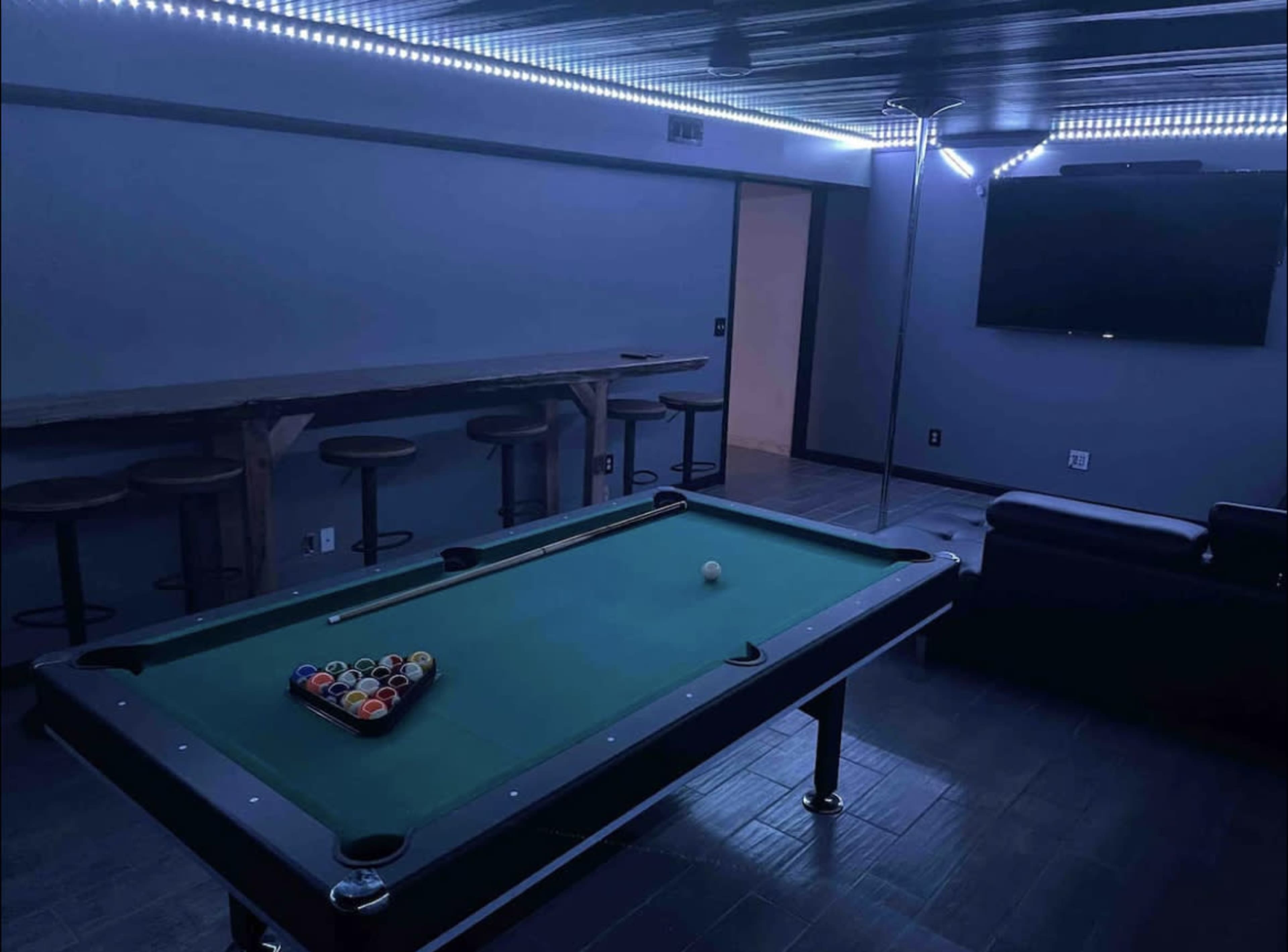 A billiards table with a triangle of Pool balls is set in a dimly lit game room, featuring a bar and lounge seating.