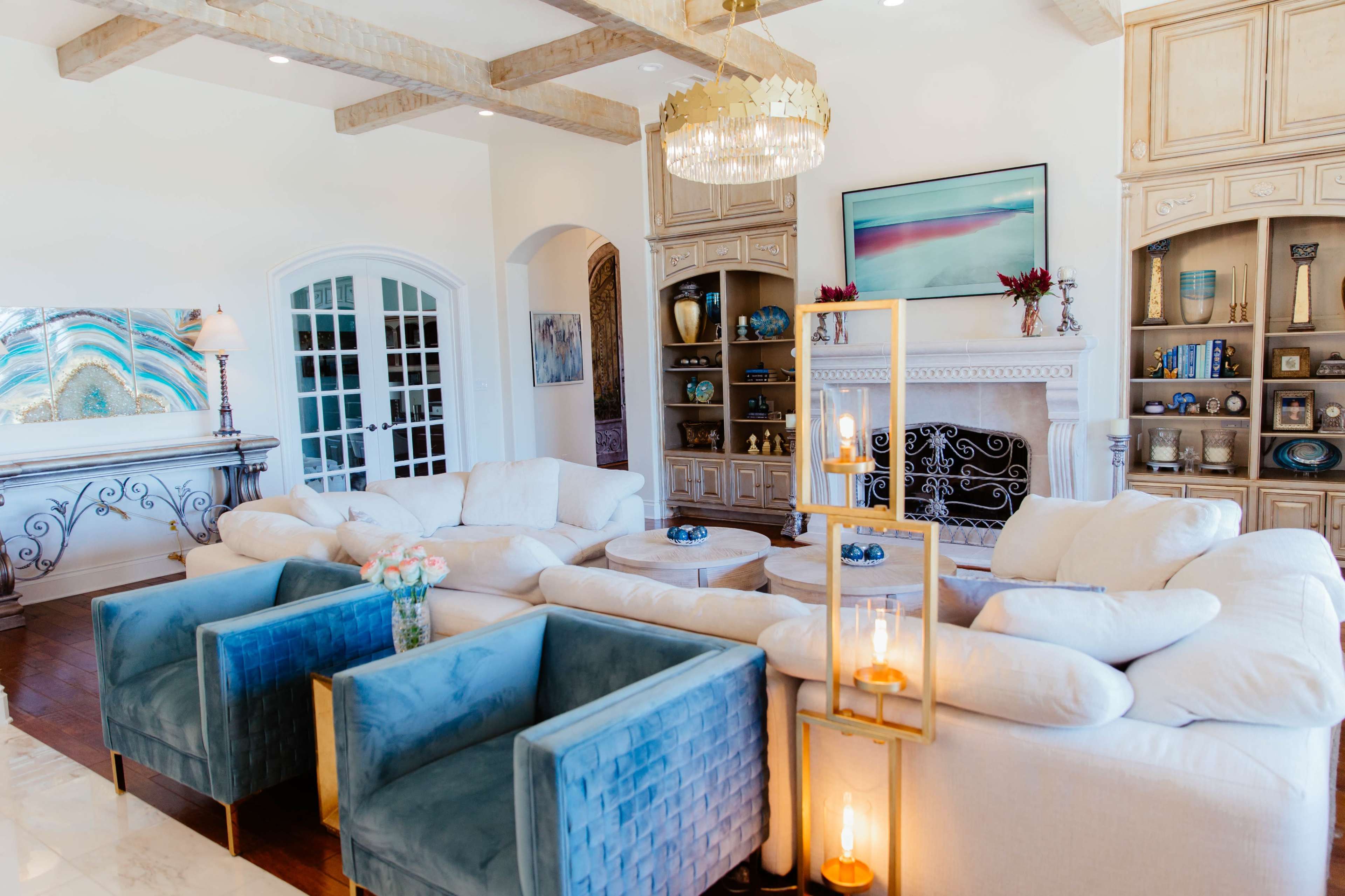 A spacious living room featuring a central seating area with a large white couch, two blue armchairs, and a decorative shelf beside a fireplace.