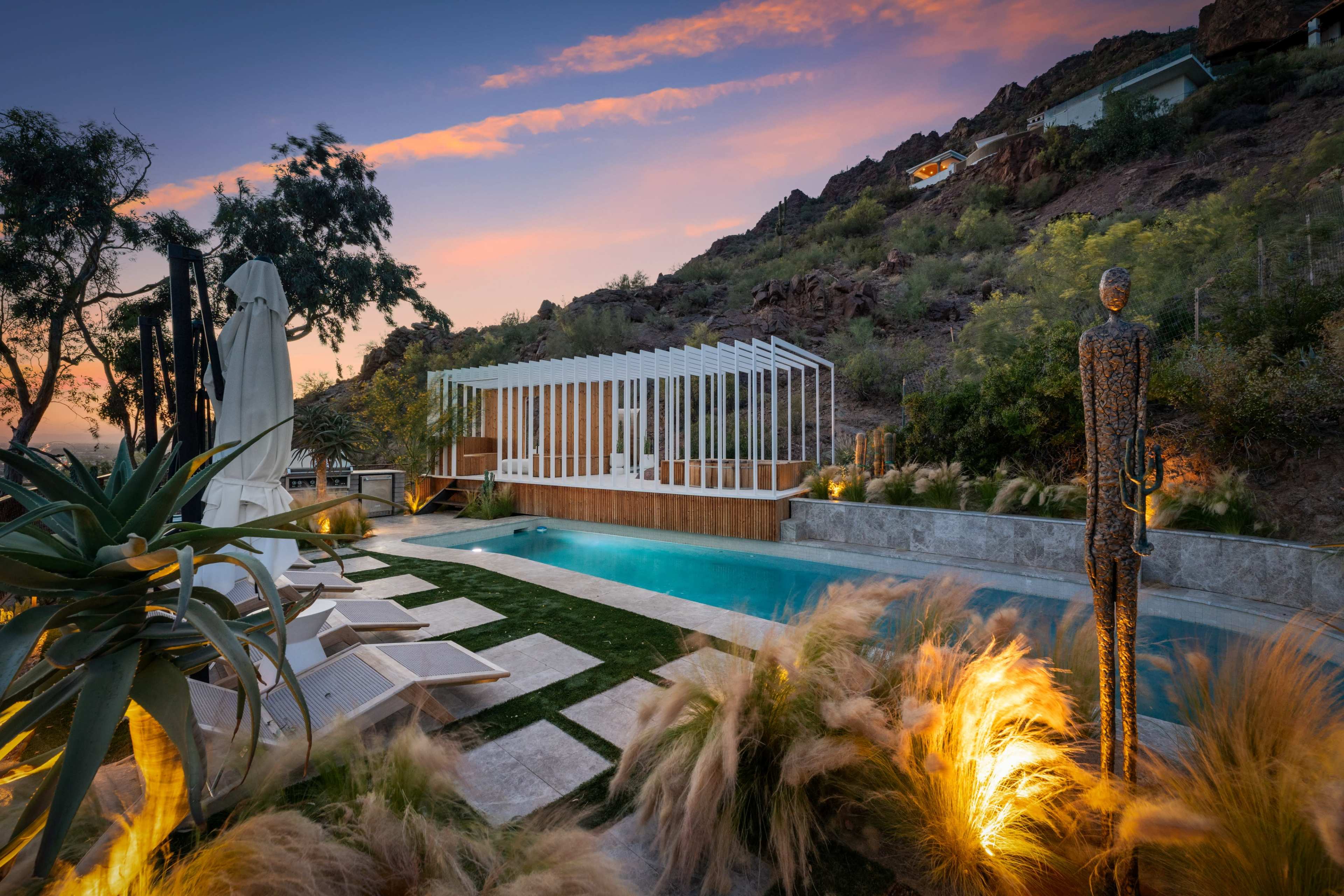 A modern outdoor pool area is set against a rugged hillside, featuring lounge chairs, a minimalist structure overhead, and landscaped greenery.