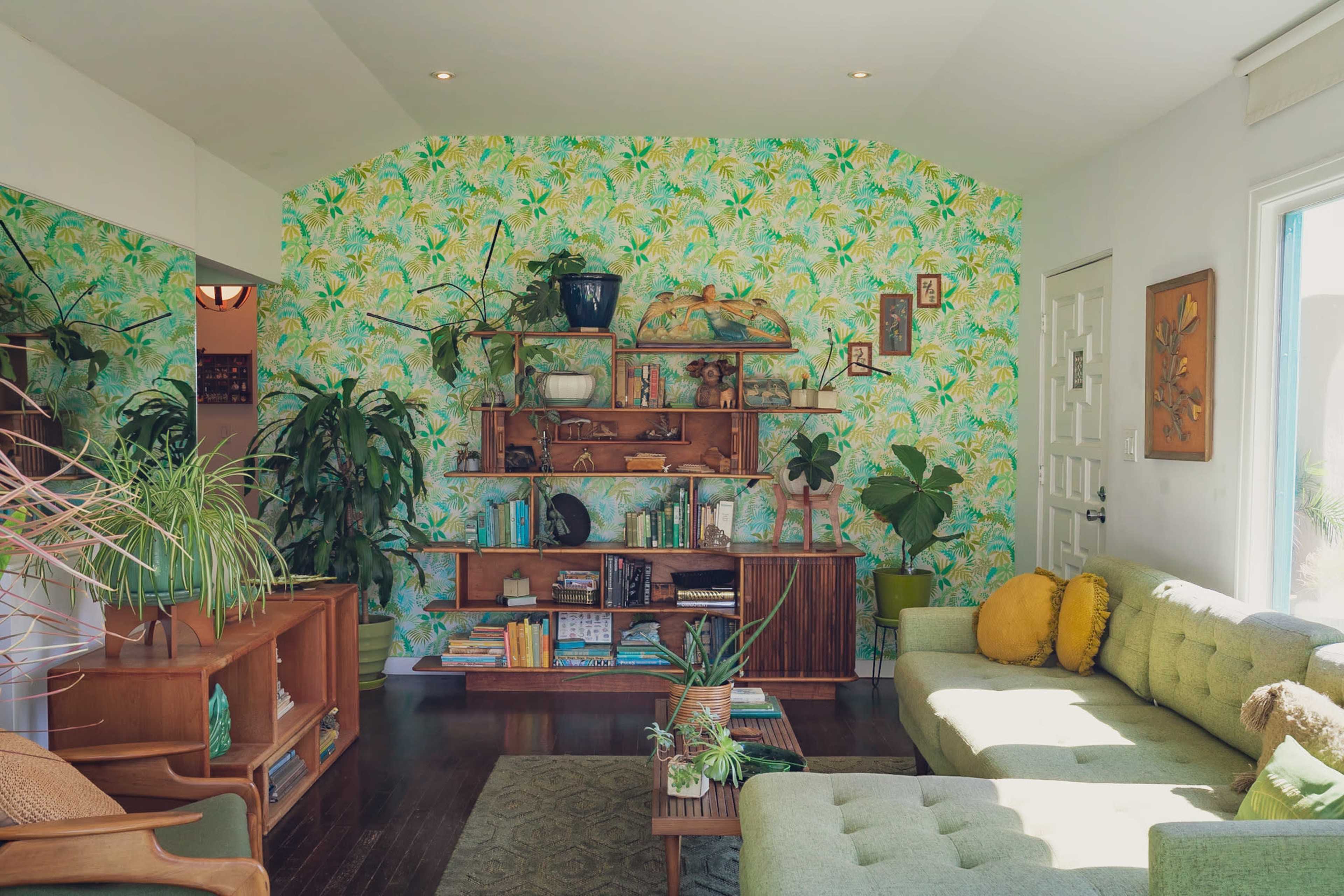 The living room features a green sectional sofa, a wooden bookshelf filled with books and plants, and a wall covered in tropical-patterned wallpaper.
