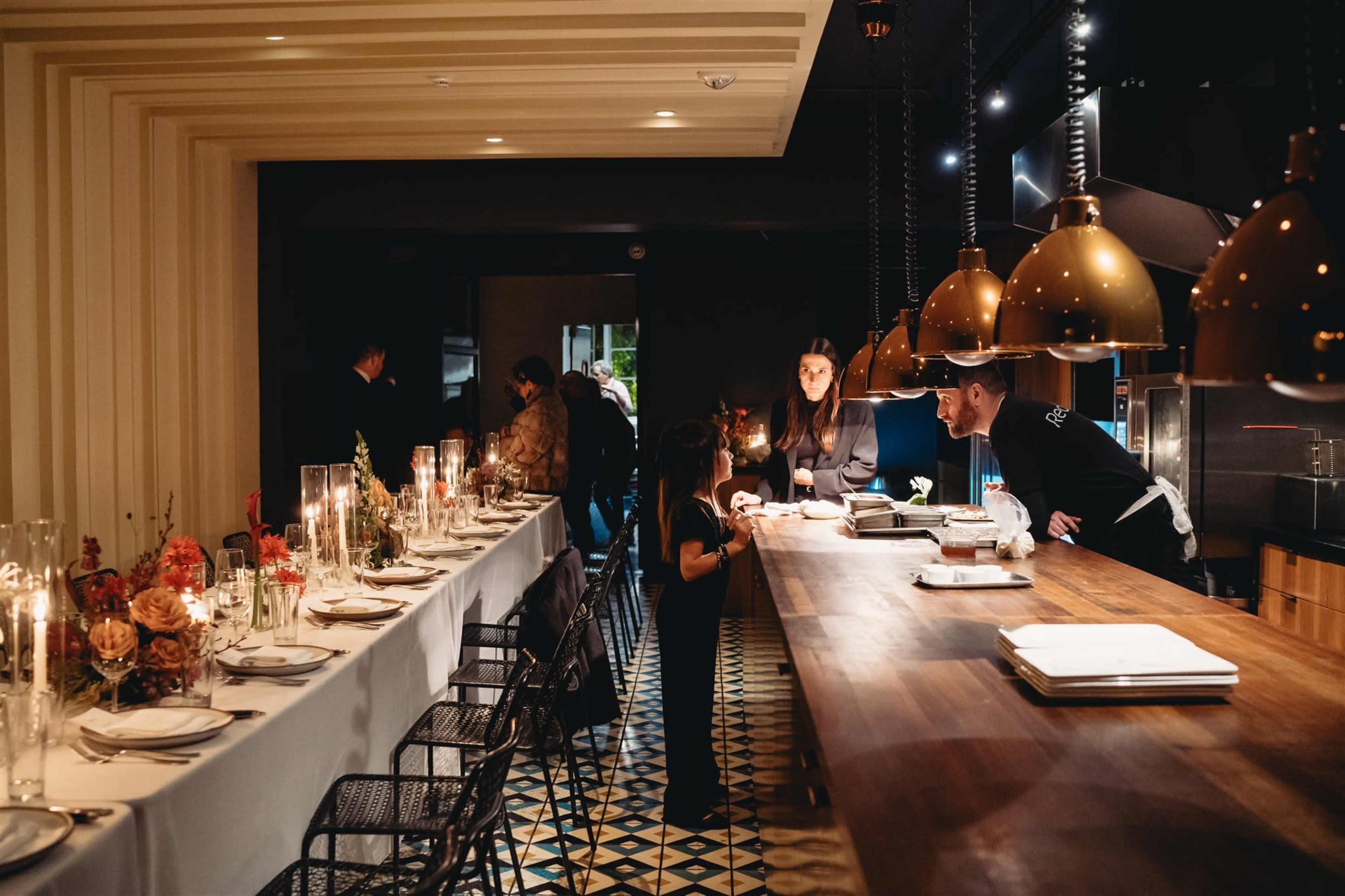 A modern restaurant interior features a long dining table set with candles, while staff prepare food at a counter under decorative pendant lights.