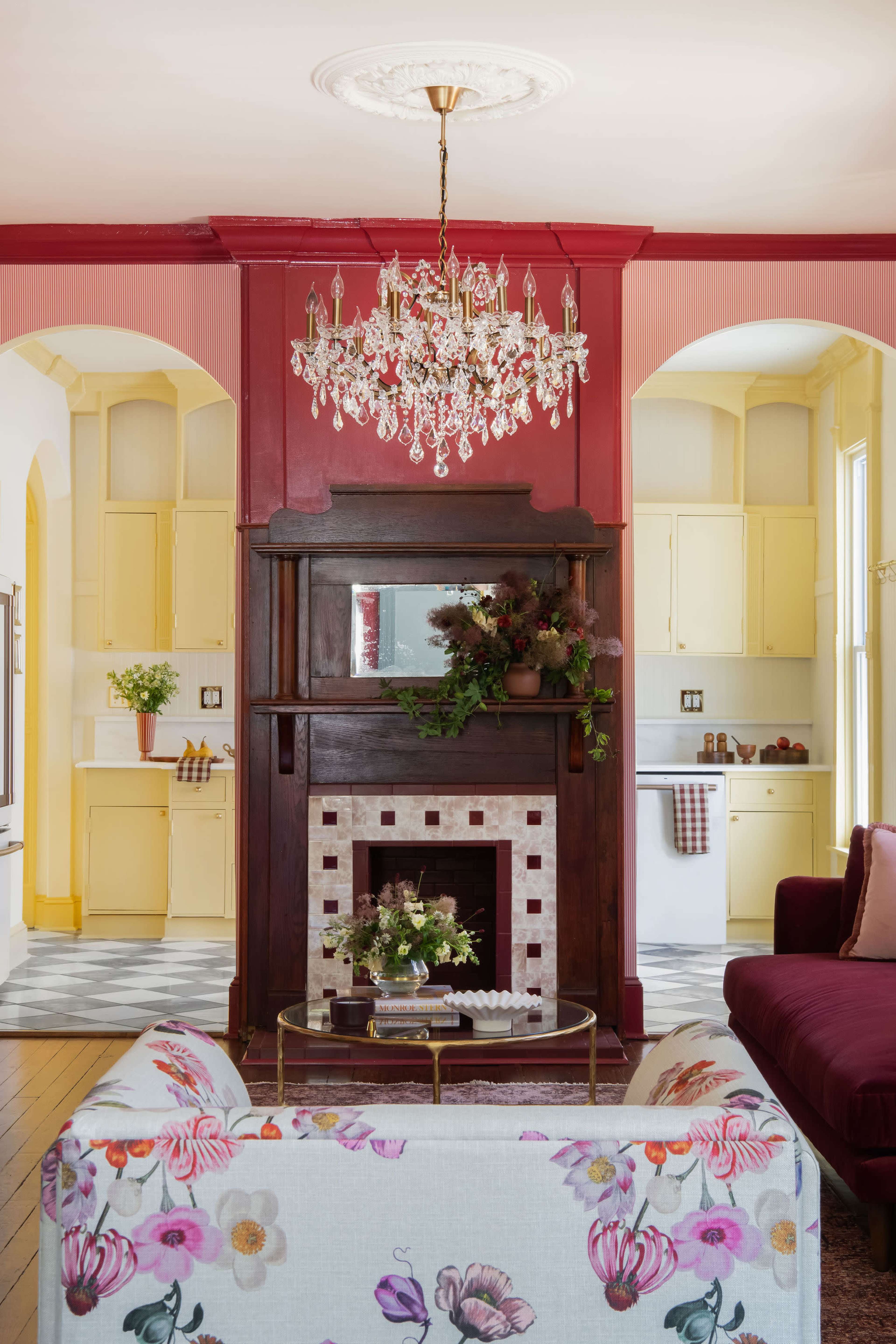 The image shows a living room with a floral-patterned sofa facing a decorative fireplace surrounded by deep red and yellow walls, and a chandelier hanging from the ceiling.