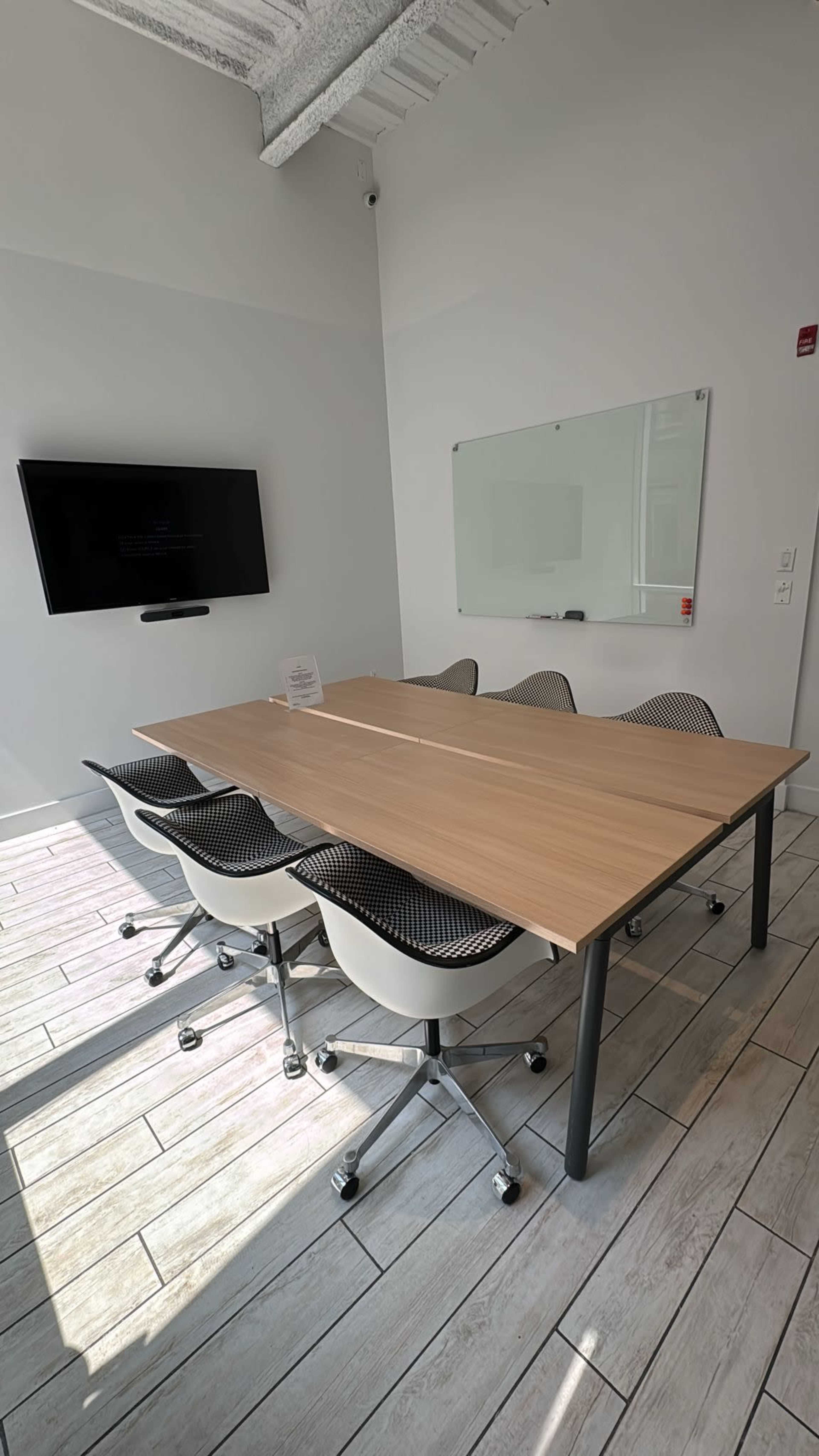 The image depicts a modern conference room with a wooden table surrounded by five swivel chairs, a television mounted on the wall, and a whiteboard beside it.