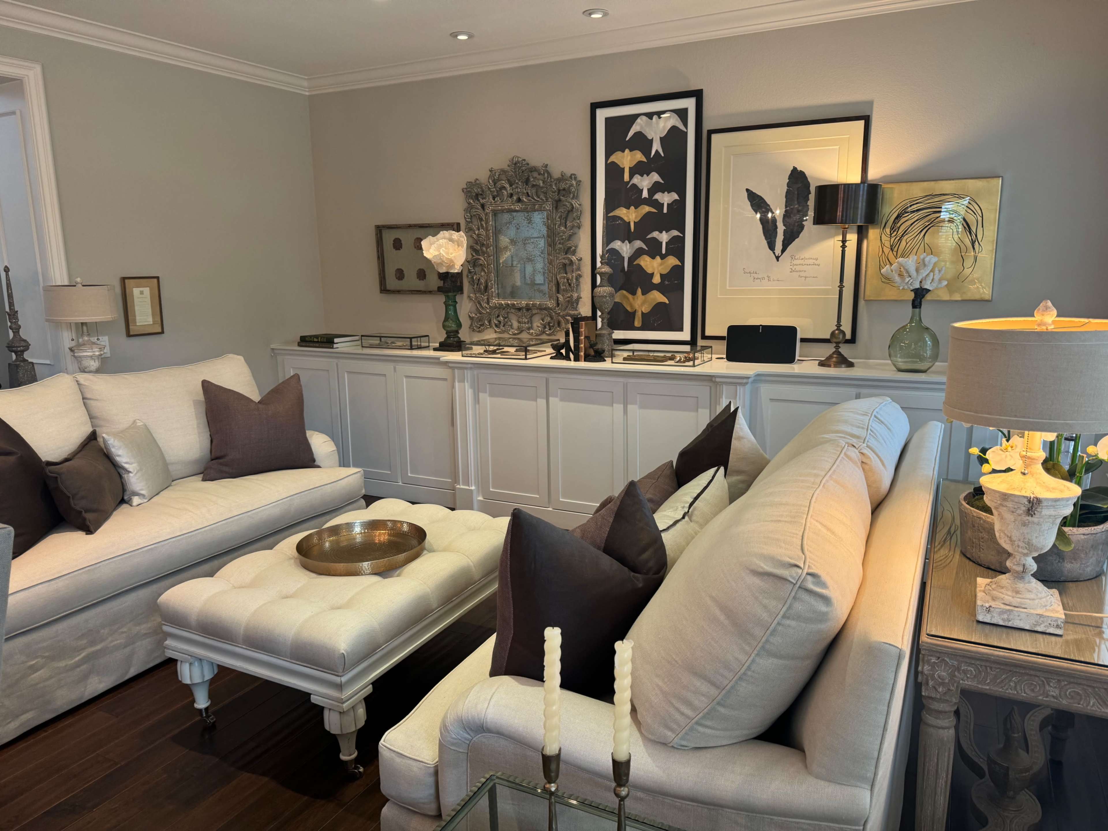 A well-decorated living room with two cream-colored sofas, a tufted ottoman, and various framed artworks displayed on a console table against a light-colored wall.