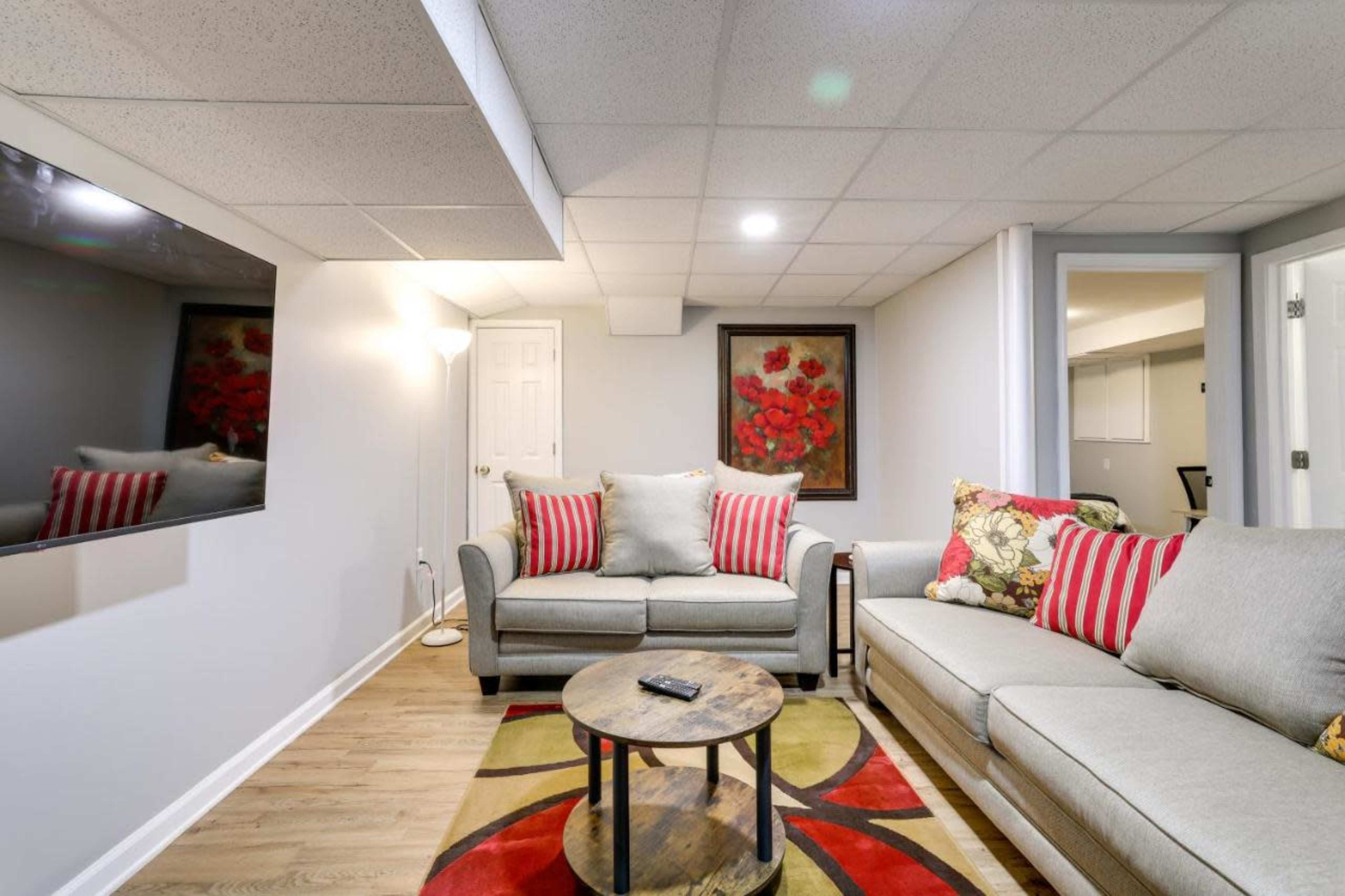 The image shows a basement living room with two sofas, a large wall-mounted television, a round coffee table, and floral artwork on the wall.