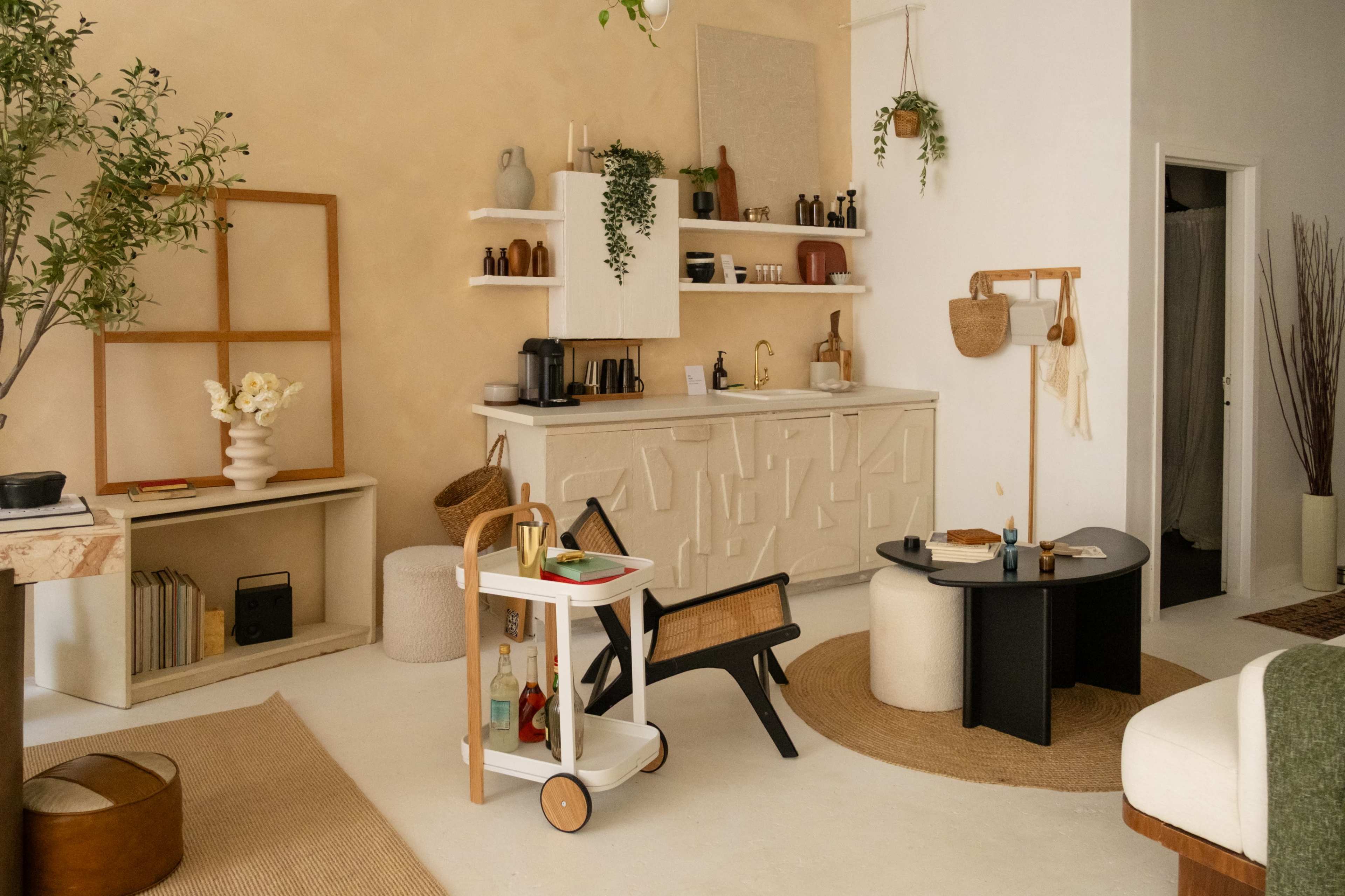 The image shows a modern, minimalist interior with light-colored walls, plants, and an arrangement of furniture including a black table, a cart, and various decorative items.