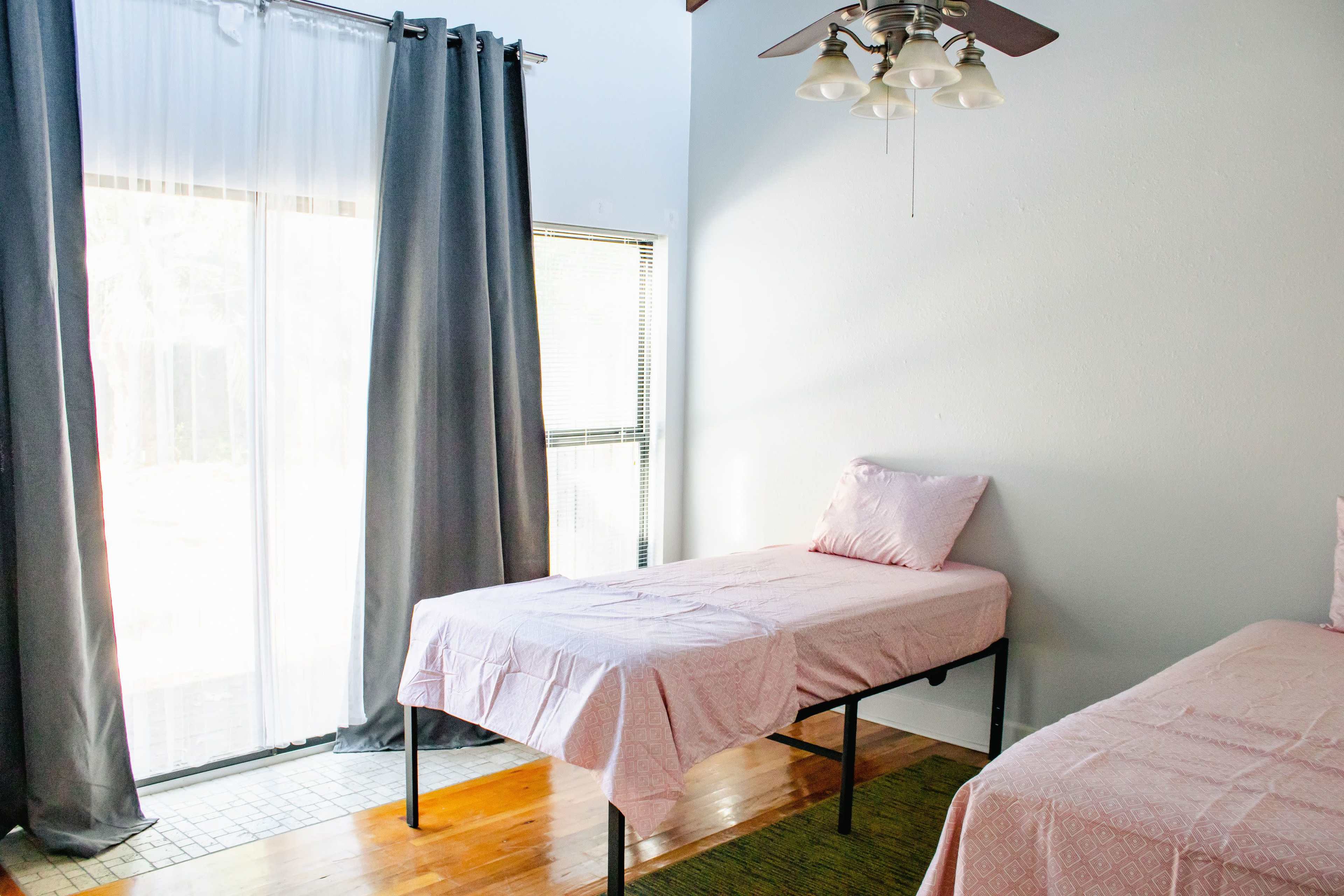 A bright room with two single beds covered in pink sheets, large windows with sheer curtains, and a ceiling fan.