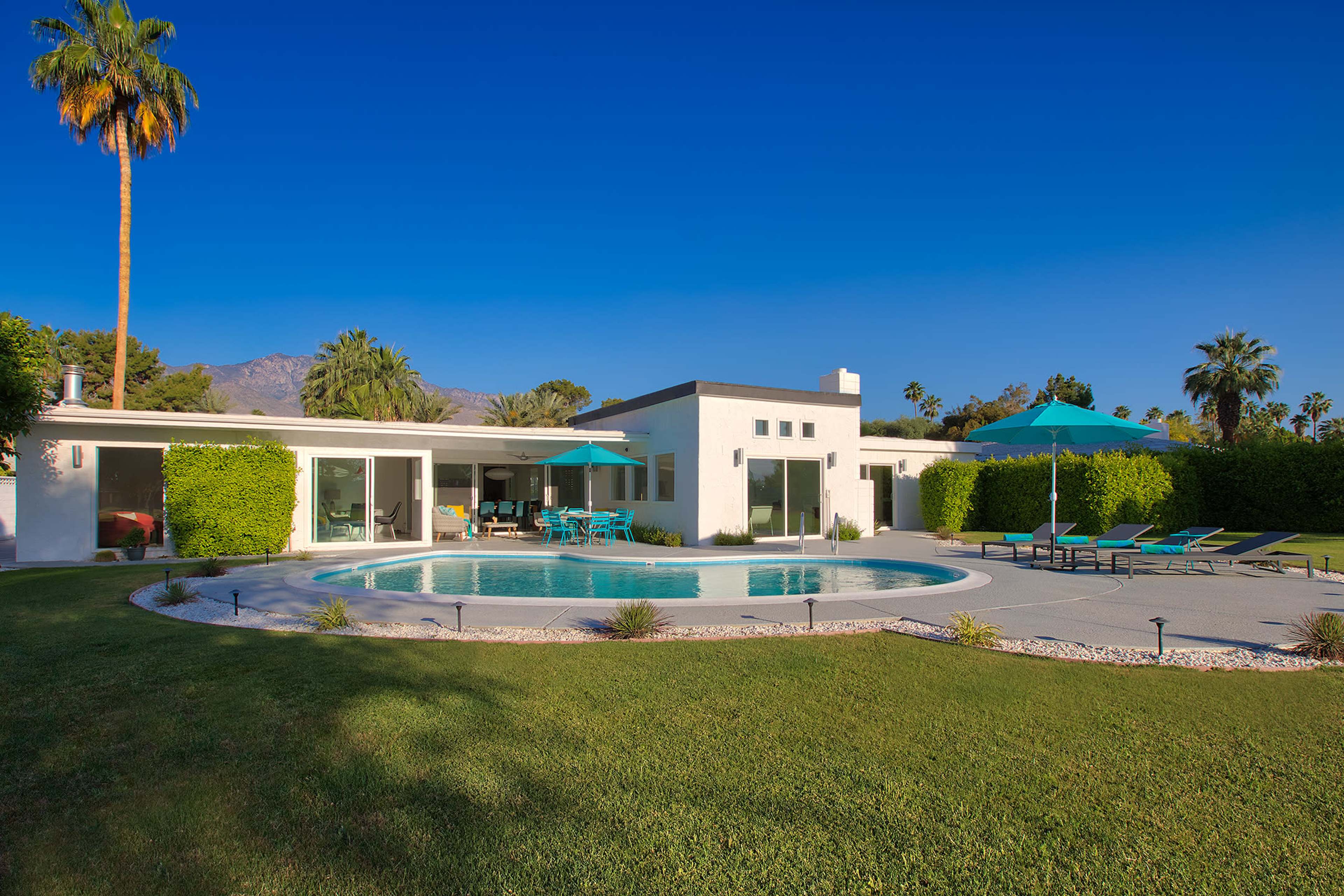 A modern residential home features a circular swimming pool surrounded by lounge chairs and palm trees against a clear blue sky.