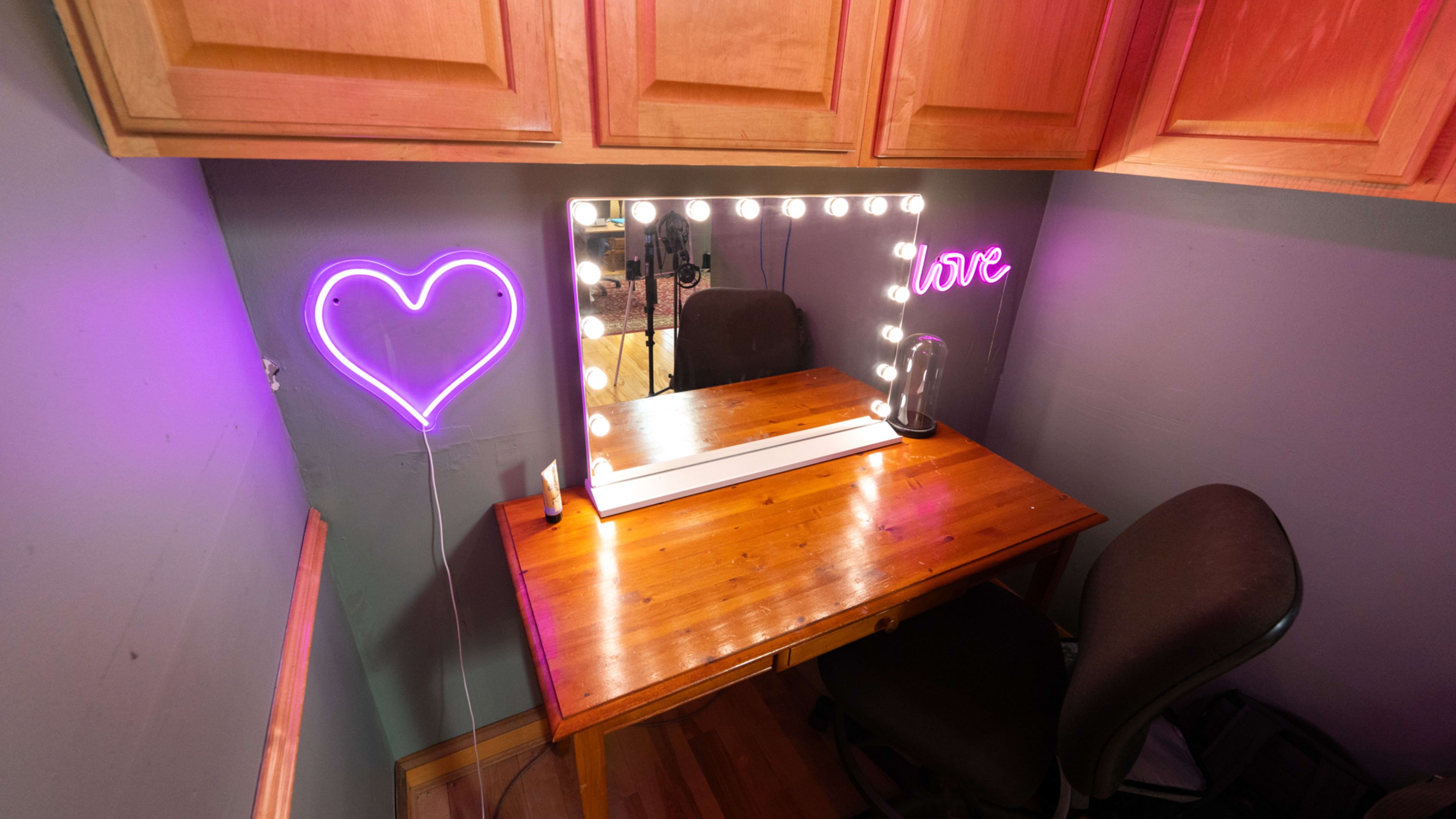 A wooden vanity table is positioned under a set of cabinets, featuring a lighted mirror and neon signs shaped like a heart and the word "love."