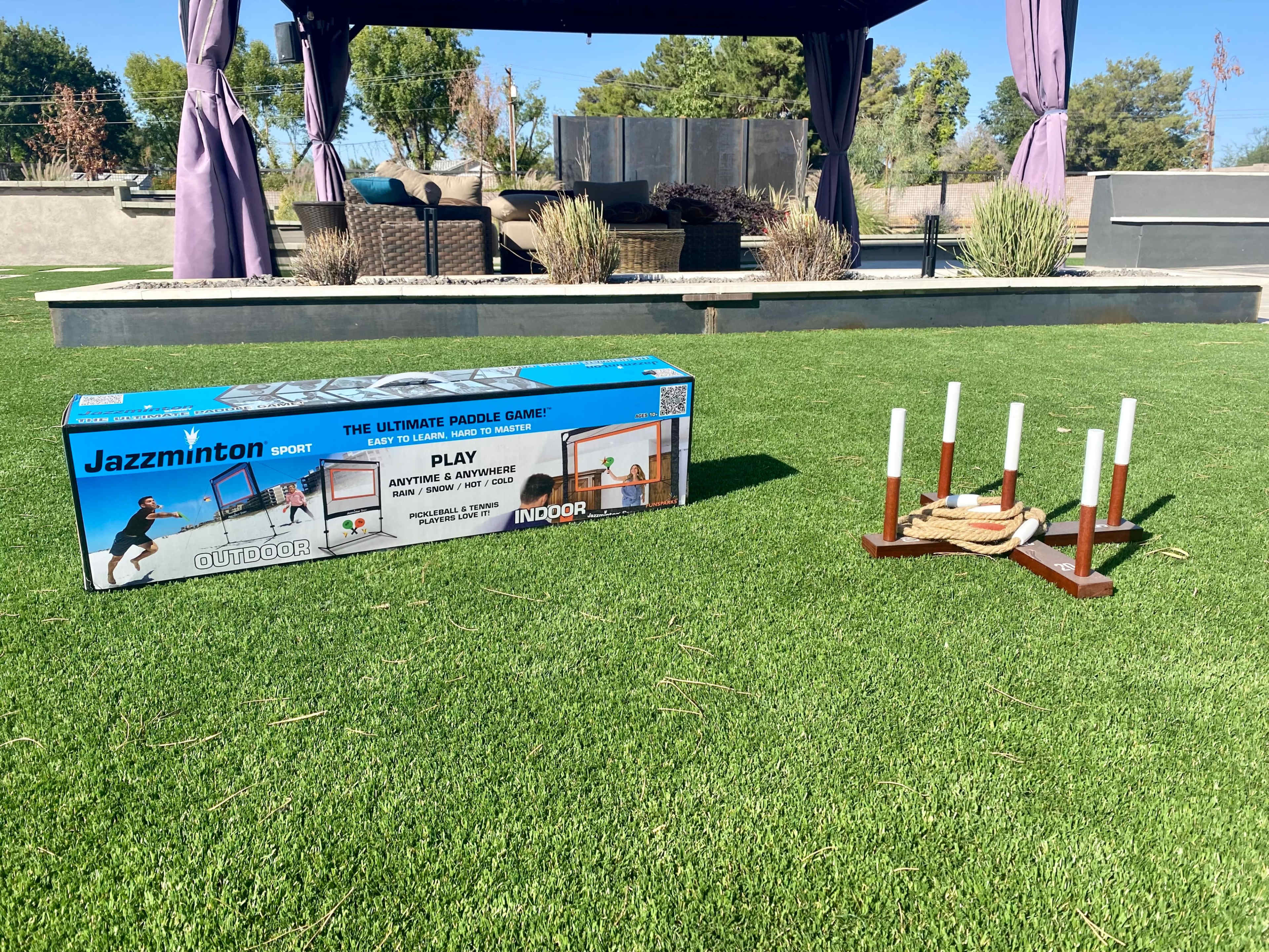 A box labeled "Jazzminton Sport" sits on green grass next to a wooden game set with vertical poles and a rope.