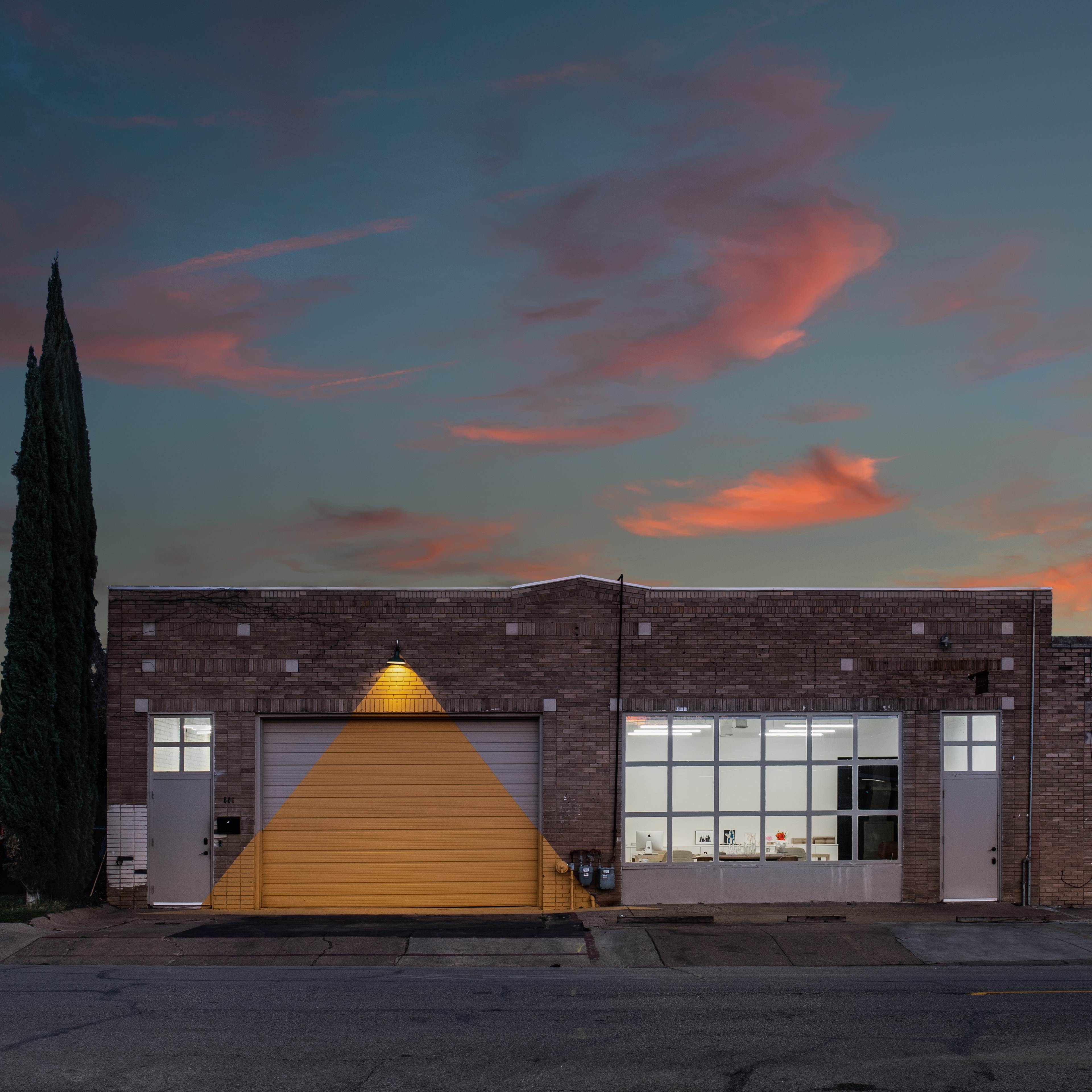 A brick building features a large yellow garage door and glass windows, set against a colorful sunset sky.