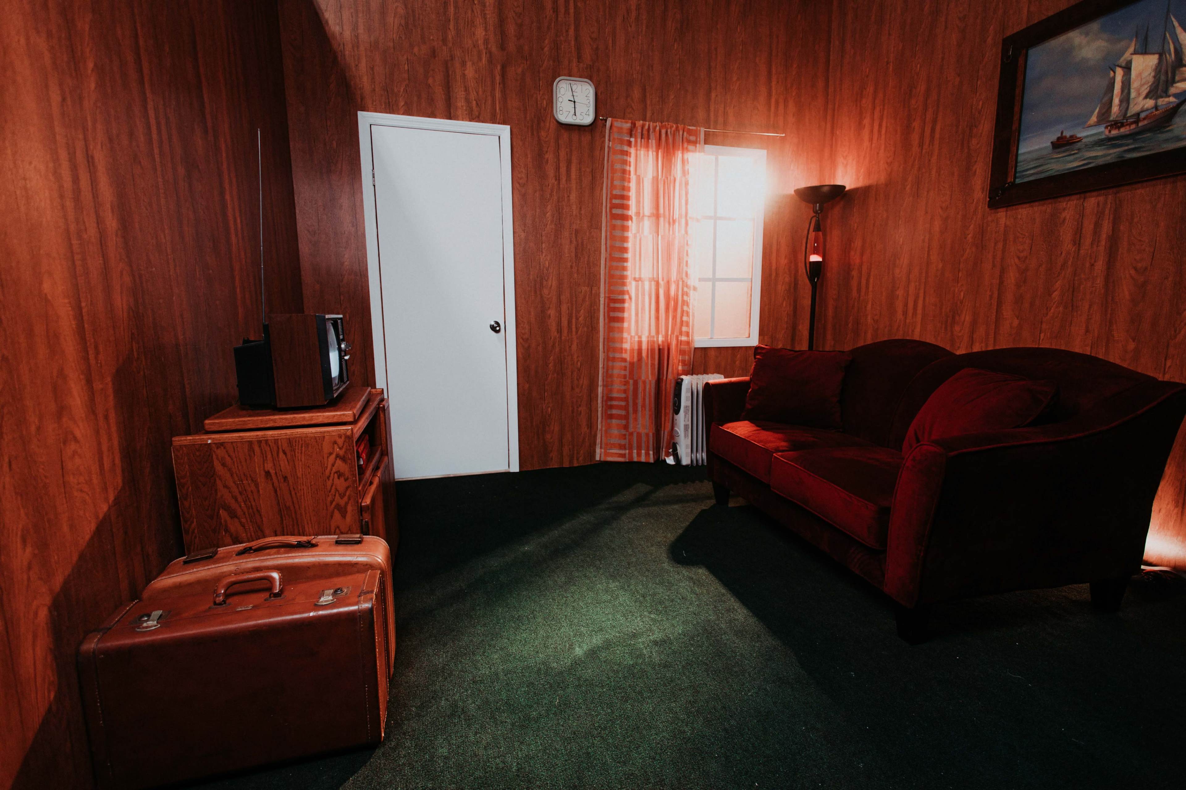 A small room with wooden walls, a red couch, an old television on a wooden cabinet, a suitcase on the green carpet, and a window with sheer curtains letting in light.