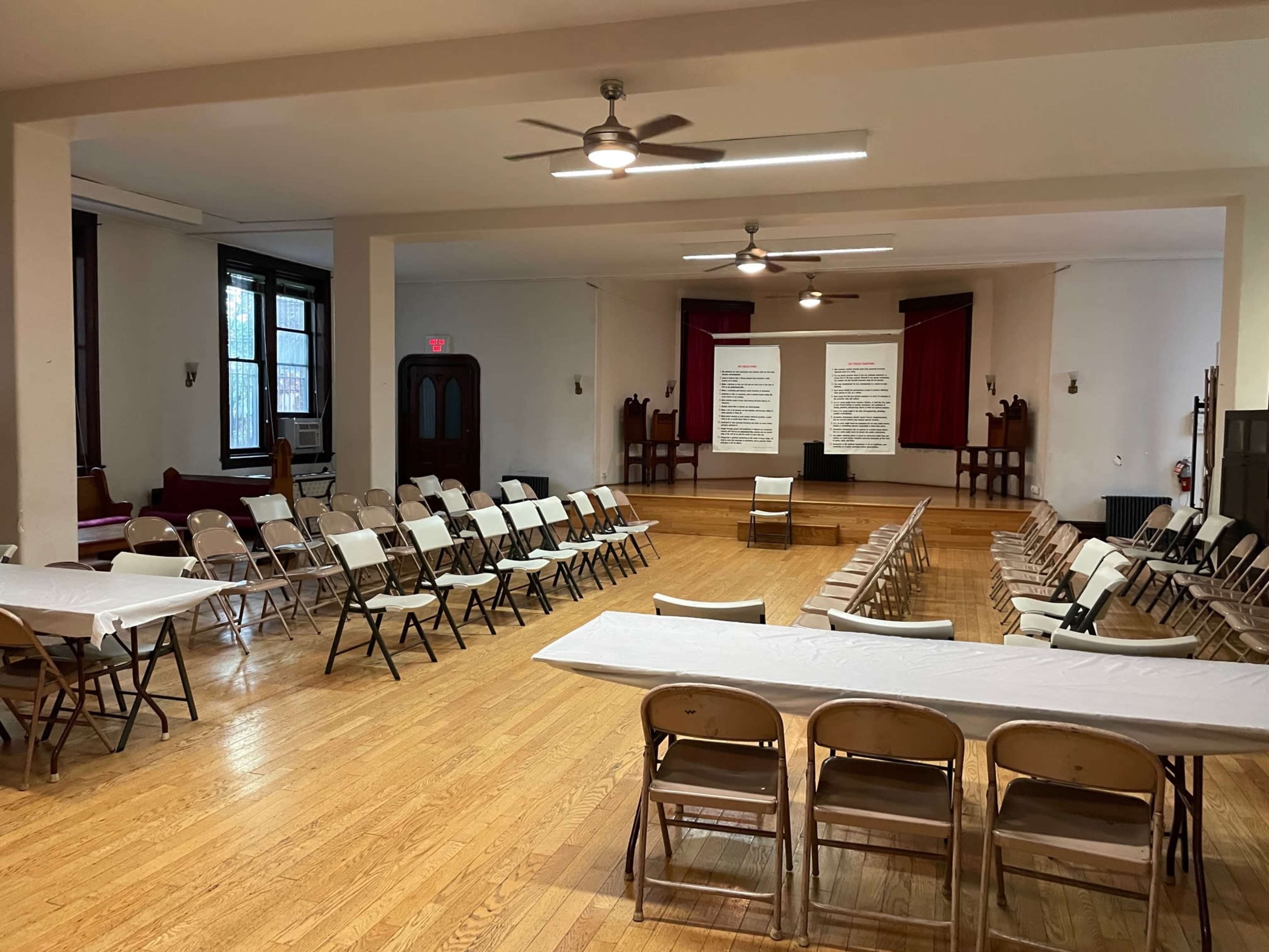 A large, empty room set up with rows of folding chairs and tables, featuring a stage area at the front with red curtains.