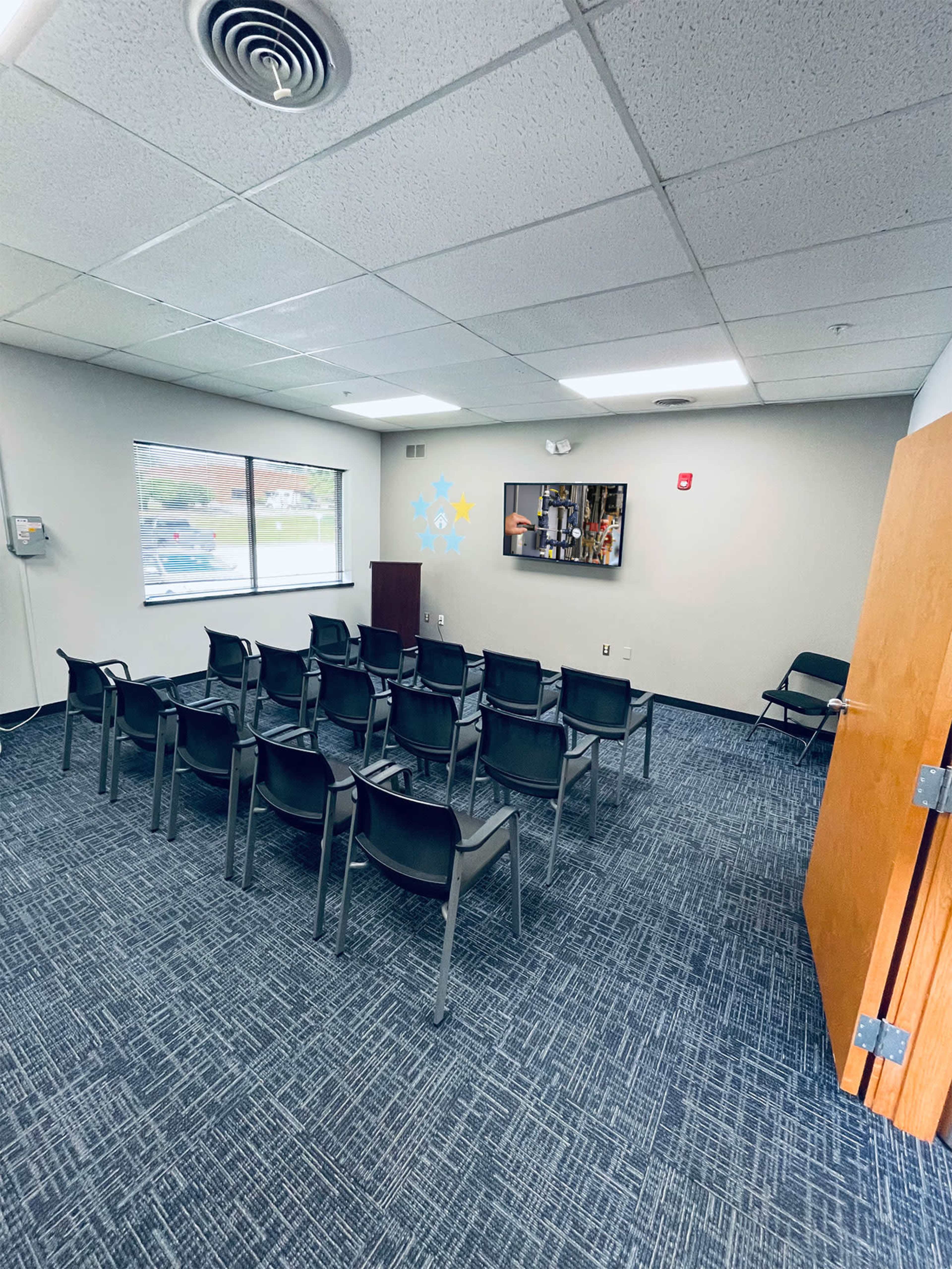 A sparsely furnished meeting room with several rows of black chairs, a small podium, and a wall-mounted display.