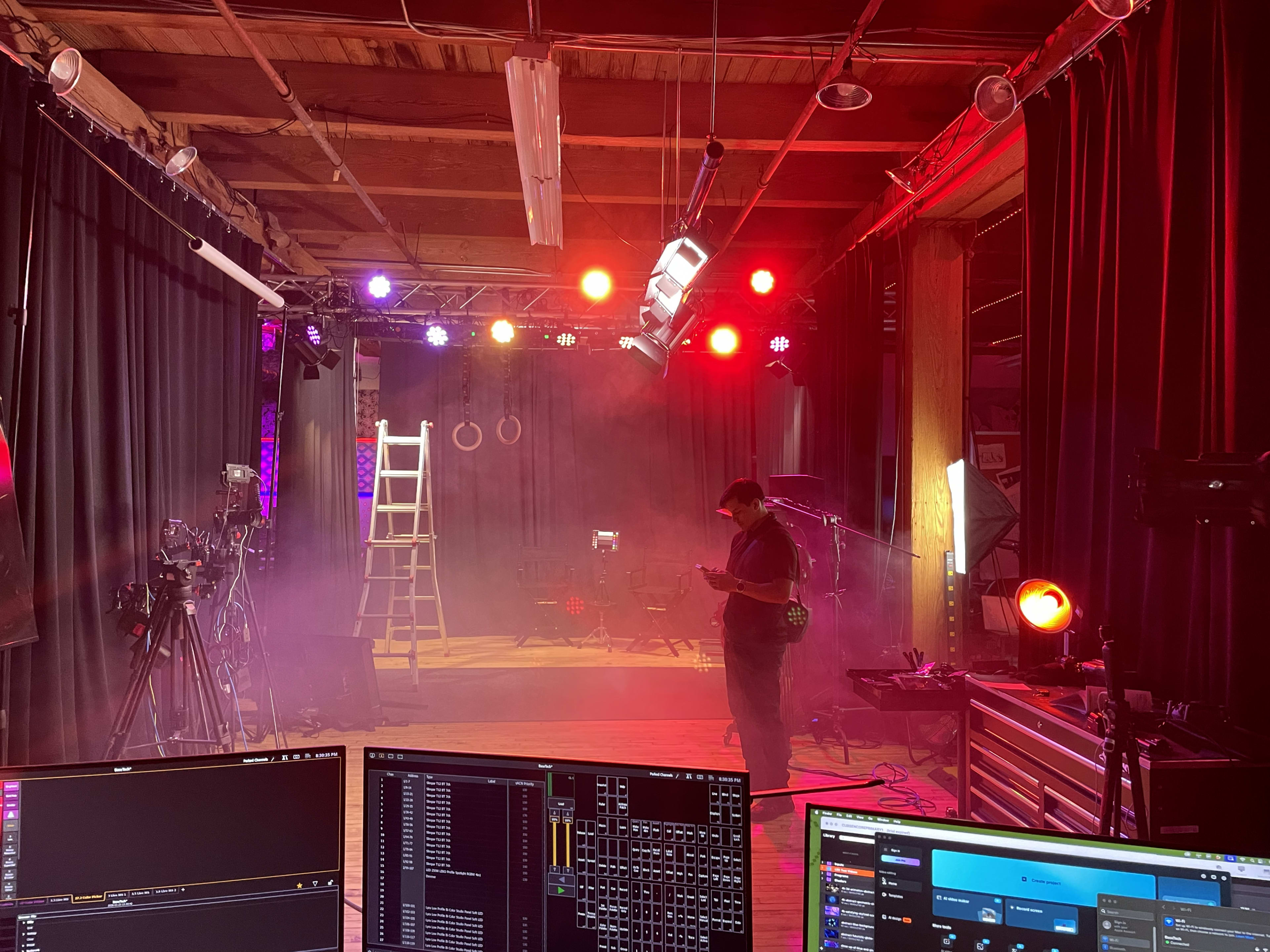 The image shows a dimly lit studio space with multicolored lights, fog, and a crew member standing near a ladder amidst filming equipment.