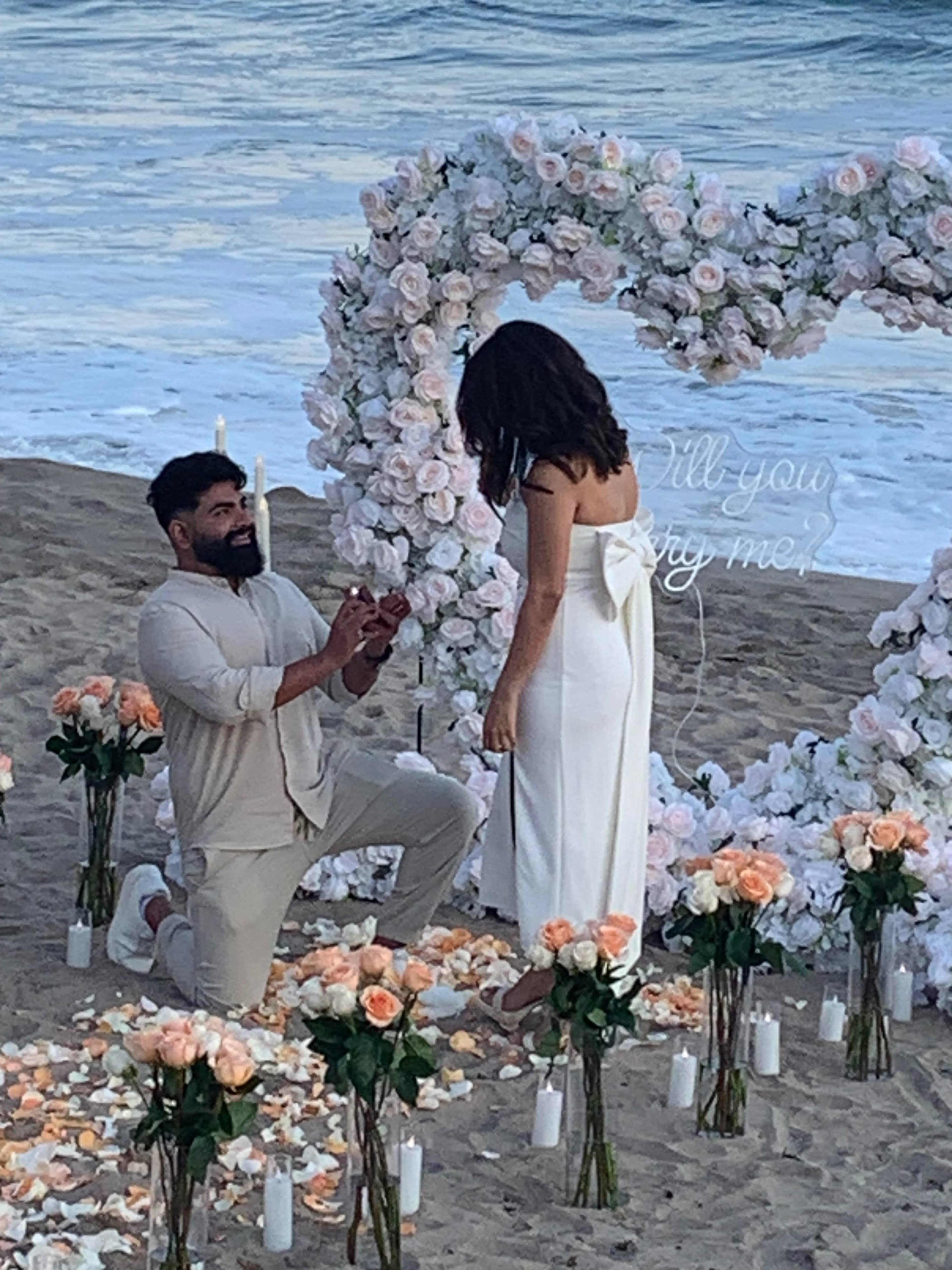 A man is proposing to a woman on the beach, kneeling in front of a floral heart arrangement with candles and rose petals scattered around them.