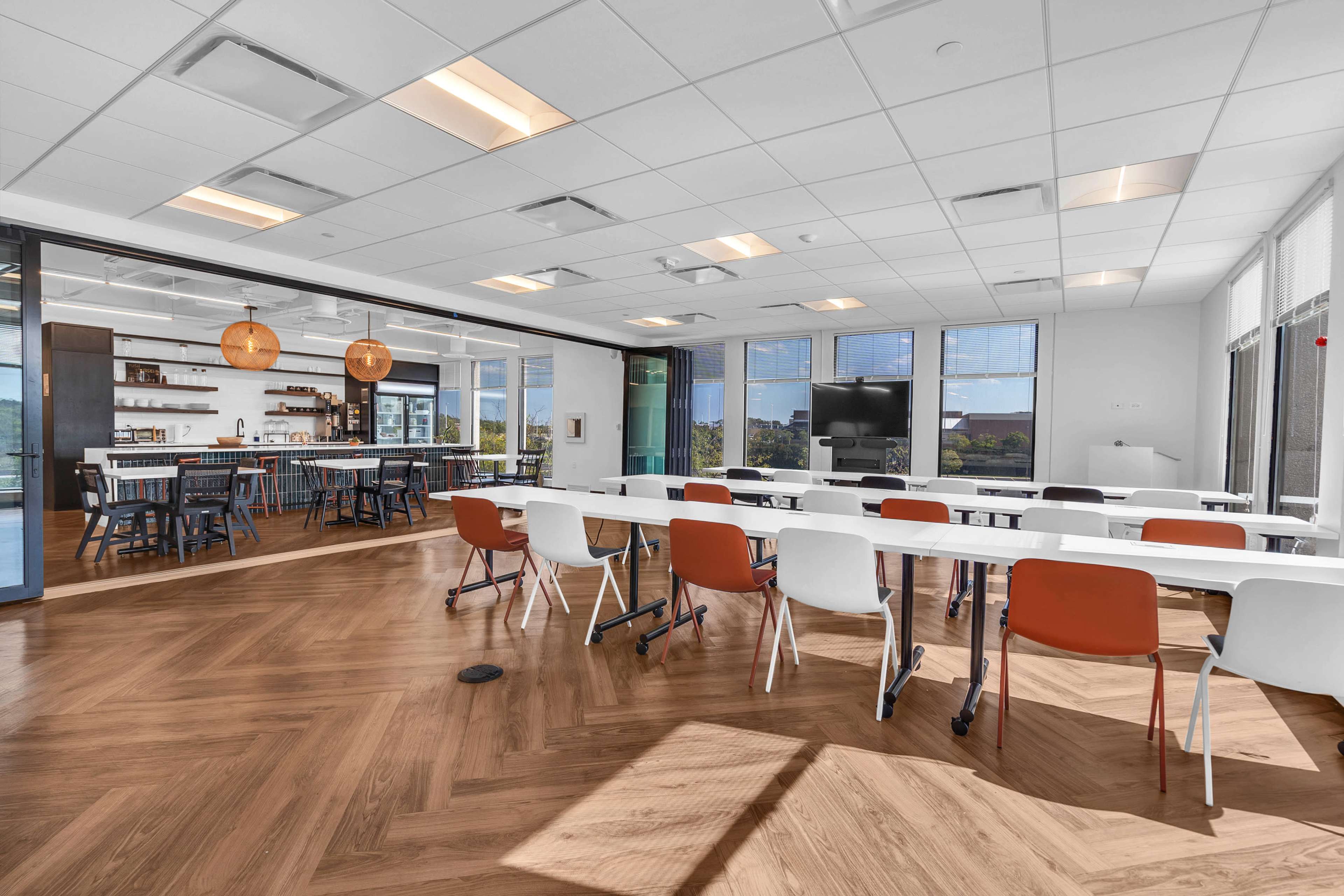 The image shows a modern, spacious classroom or meeting room with white tables, colorful chairs, large windows, and a kitchenette area.