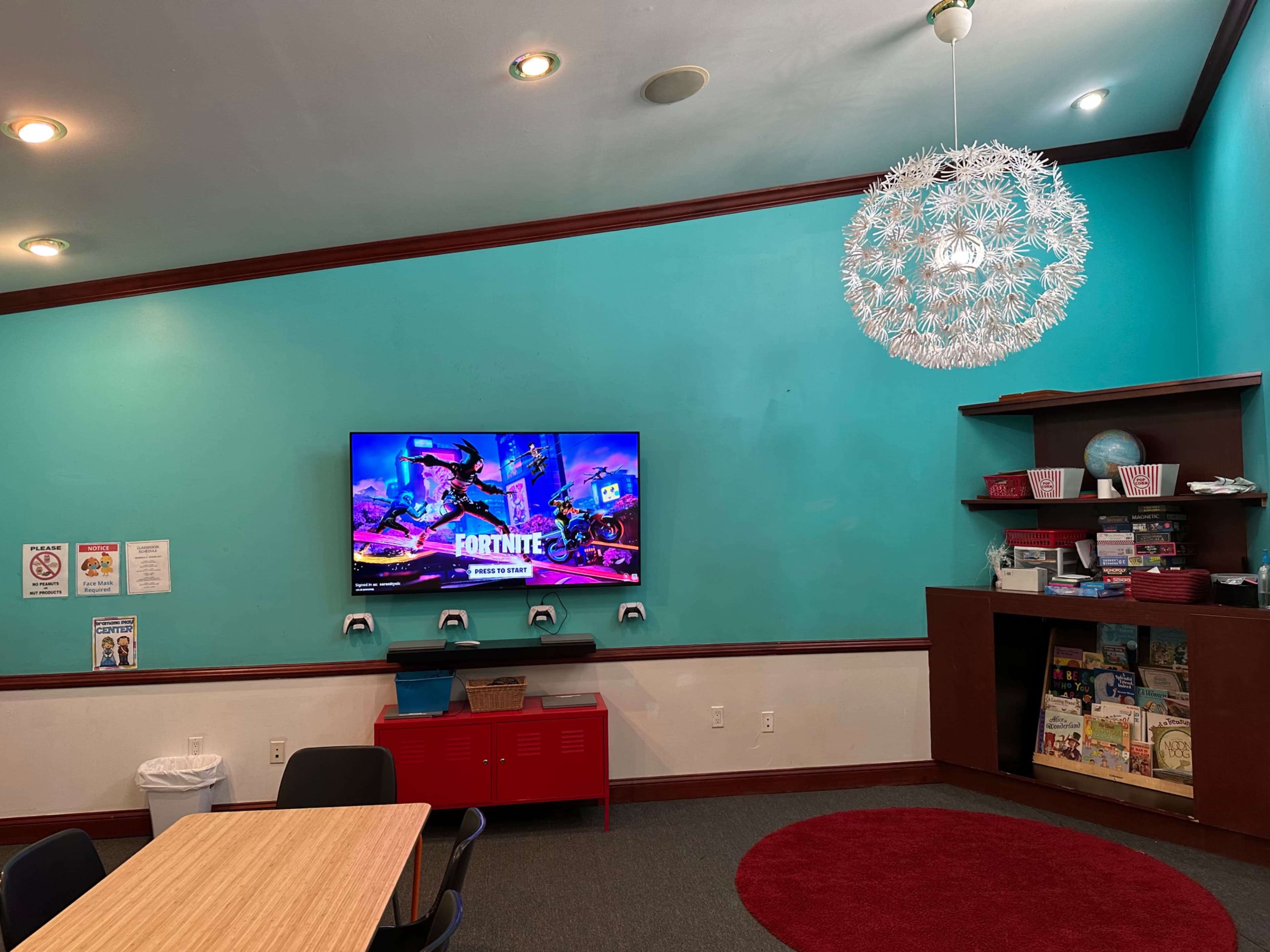 A colorful room with a television displaying a "Fortnite" screen, a circular rug on the floor, a bookshelf filled with books, and a modern light fixture hanging from the ceiling.