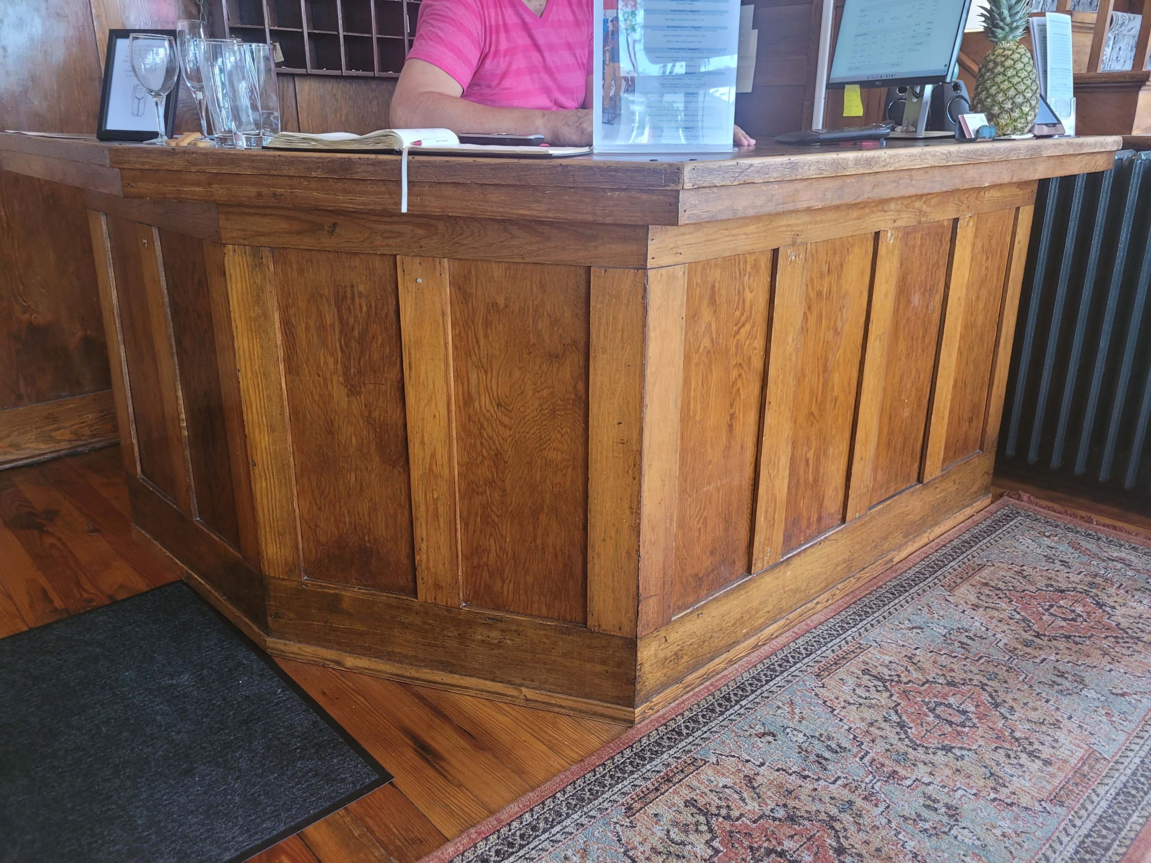 A wooden reception desk with a computer monitor and a glass of water on it, alongside a patterned rug and a black mat on the floor.