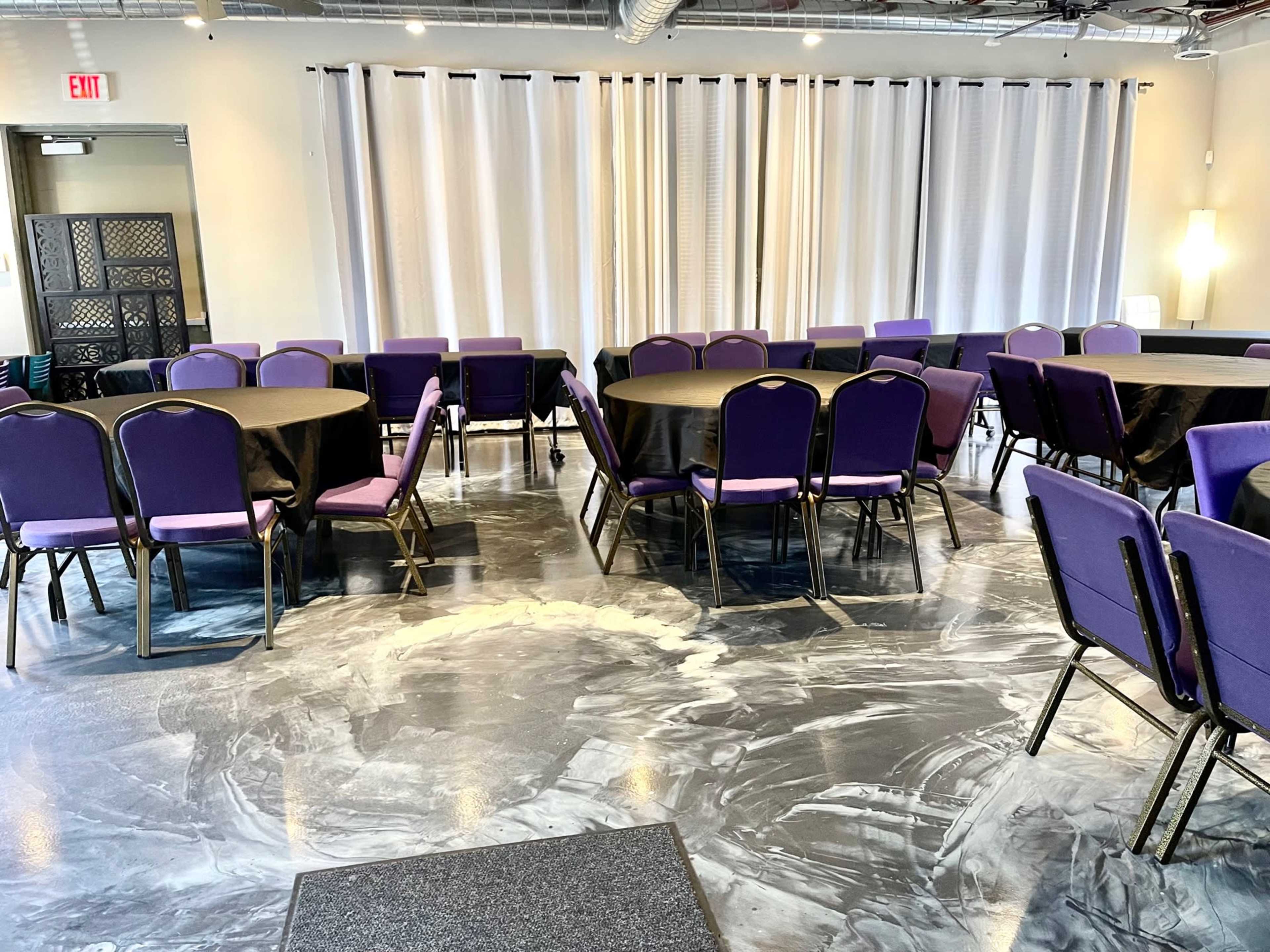 A room with several round tables covered in black tablecloths and surrounded by purple chairs, set against a backdrop of white curtains and a polished floor.
