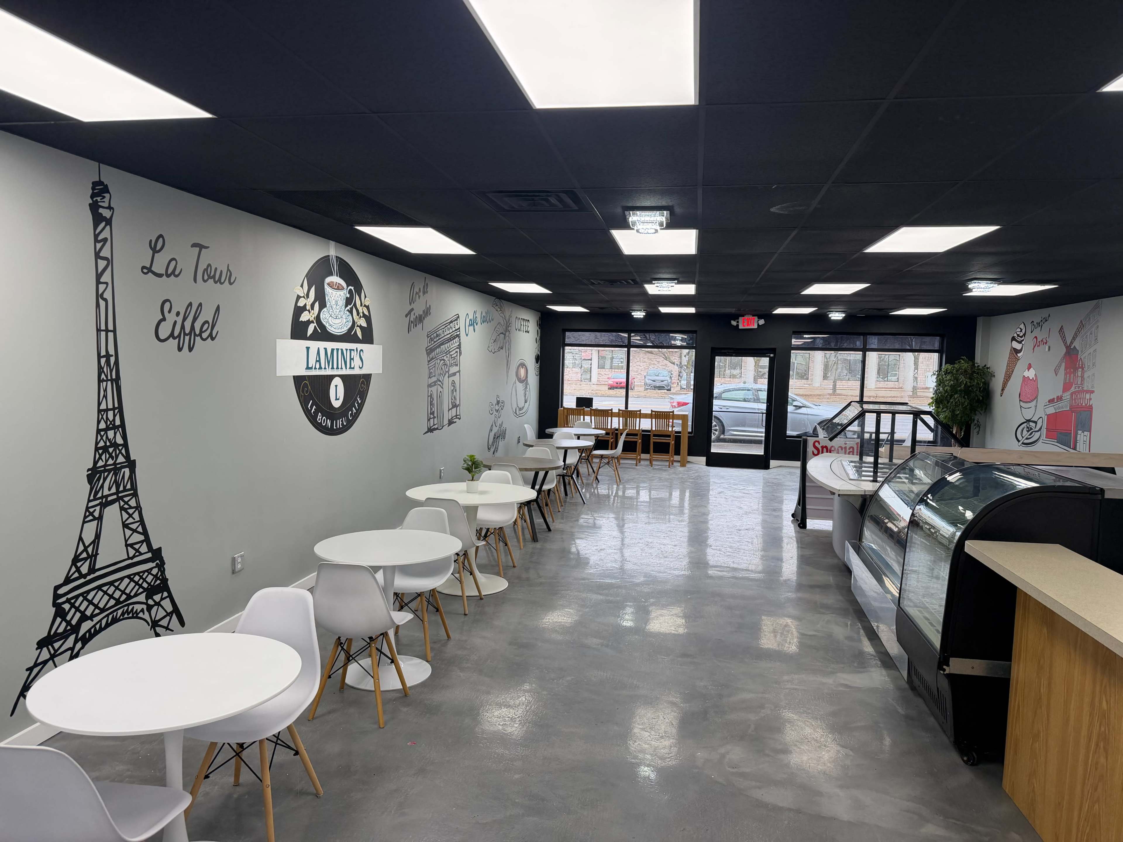 The image shows a modern café interior with white tables and chairs, a glass display case, and wall art featuring the Eiffel Tower and café branding.