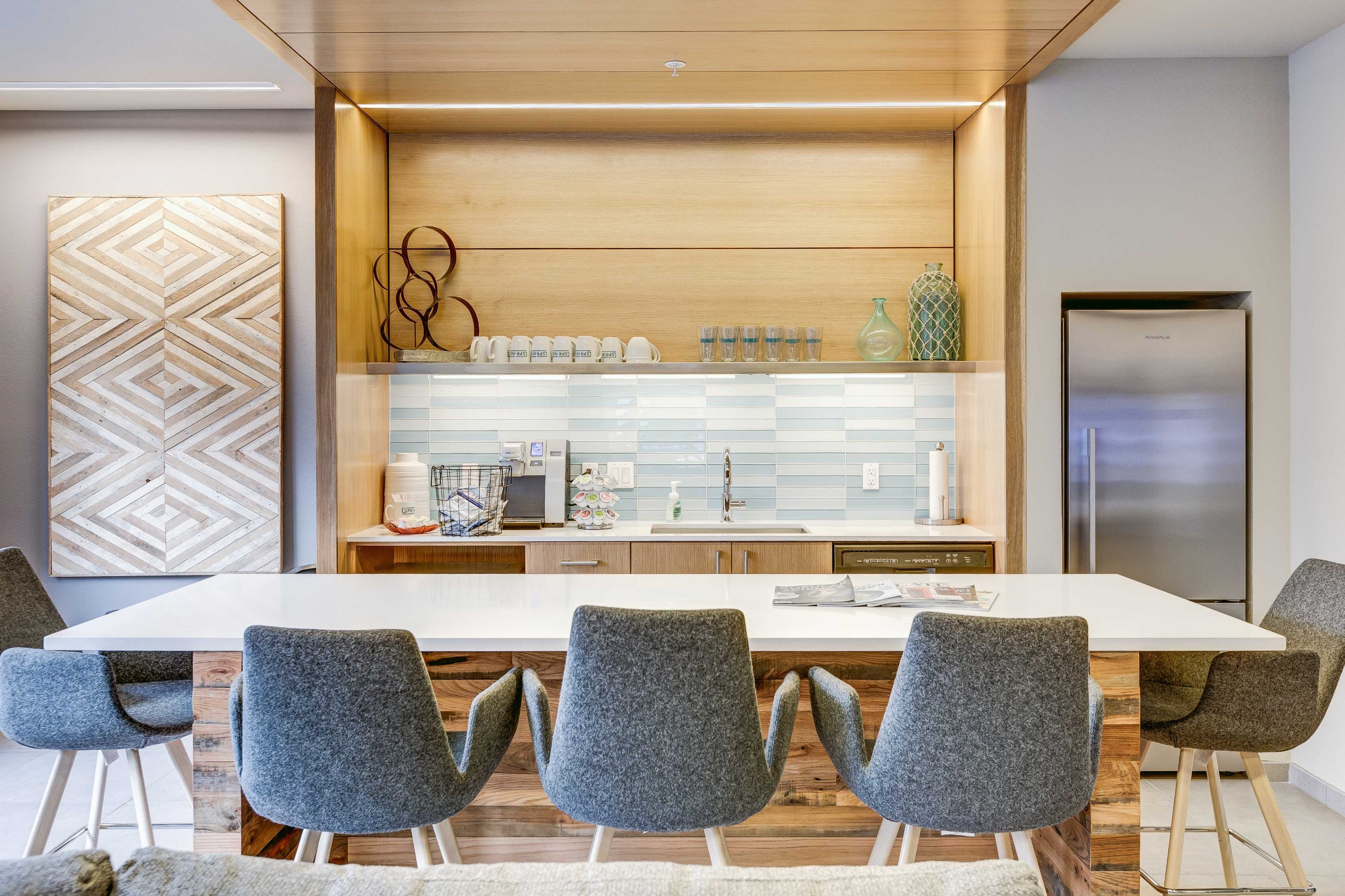 A modern kitchen bar area with a white countertop, gray upholstered chairs, wooden cabinetry, and a backsplash featuring light blue tiles.