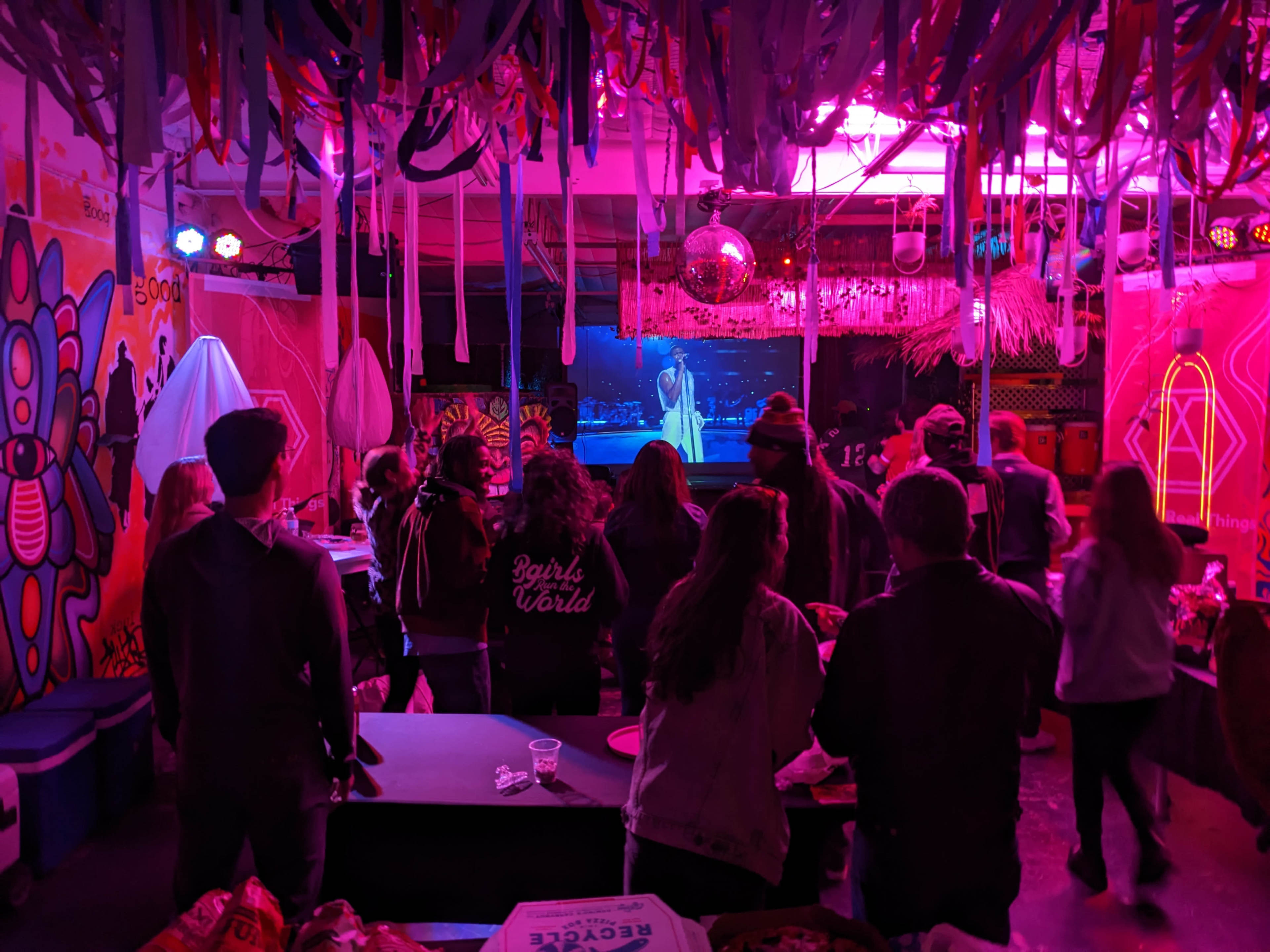 A group of people watches a performer on stage in a colorful venue adorned with streamers and vibrant decorations.