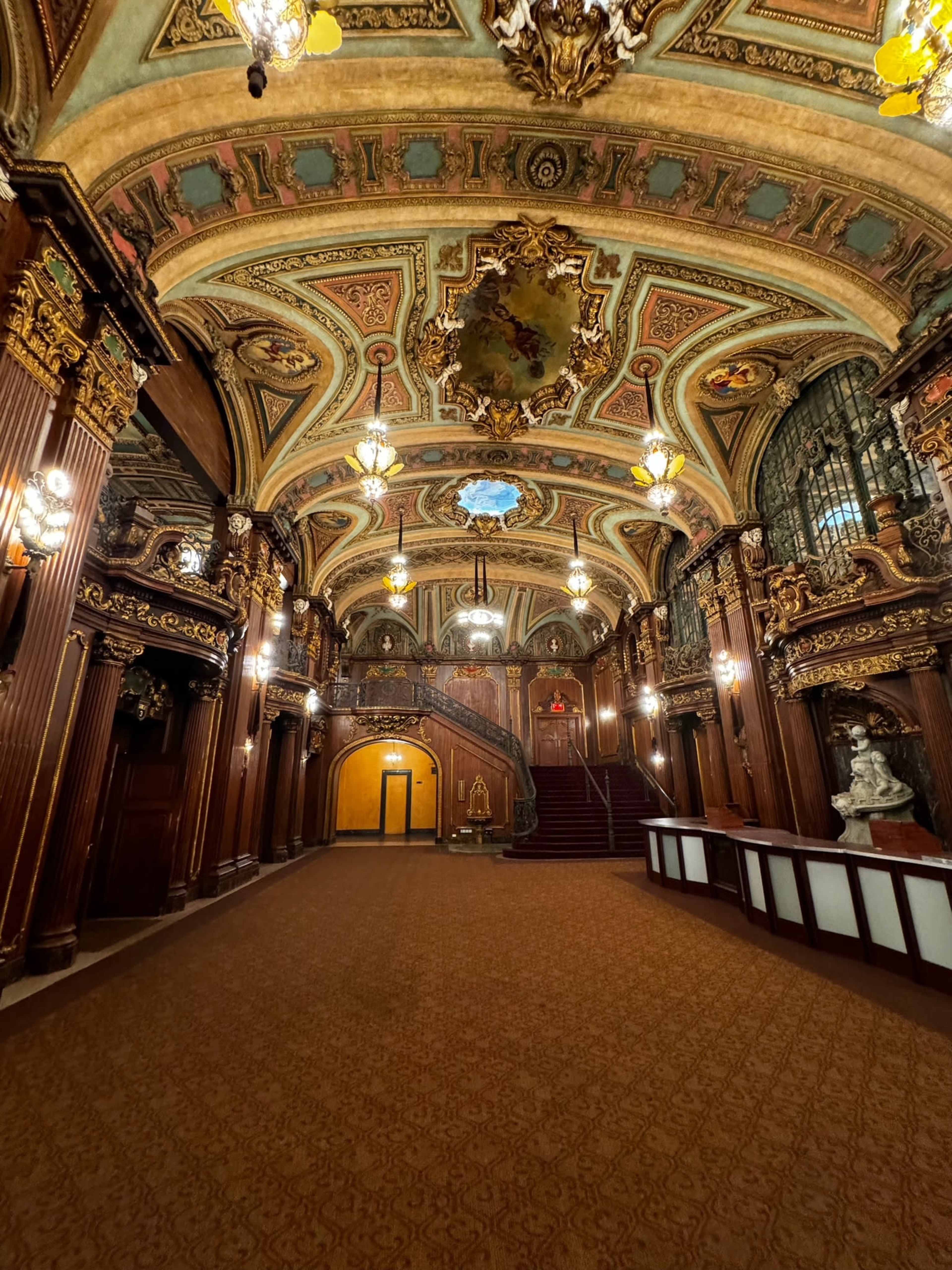 The image shows an ornate, elegantly designed interior of a historic building with decorative ceilings, wooden paneling, and a grand staircase.