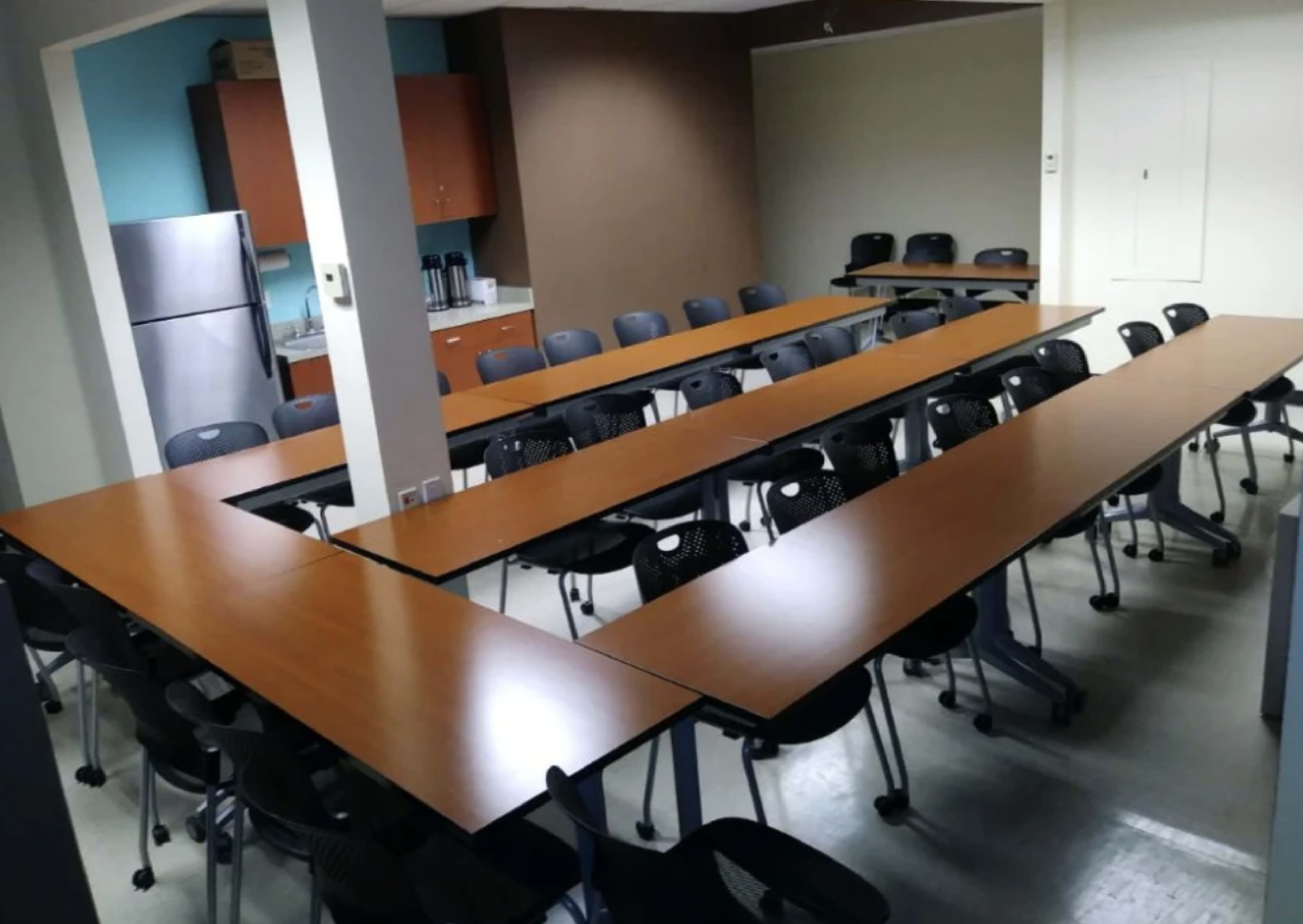 A conference room is set up with multiple long tables arranged in a U-shape, surrounded by chairs, and equipped with a kitchenette in the background.