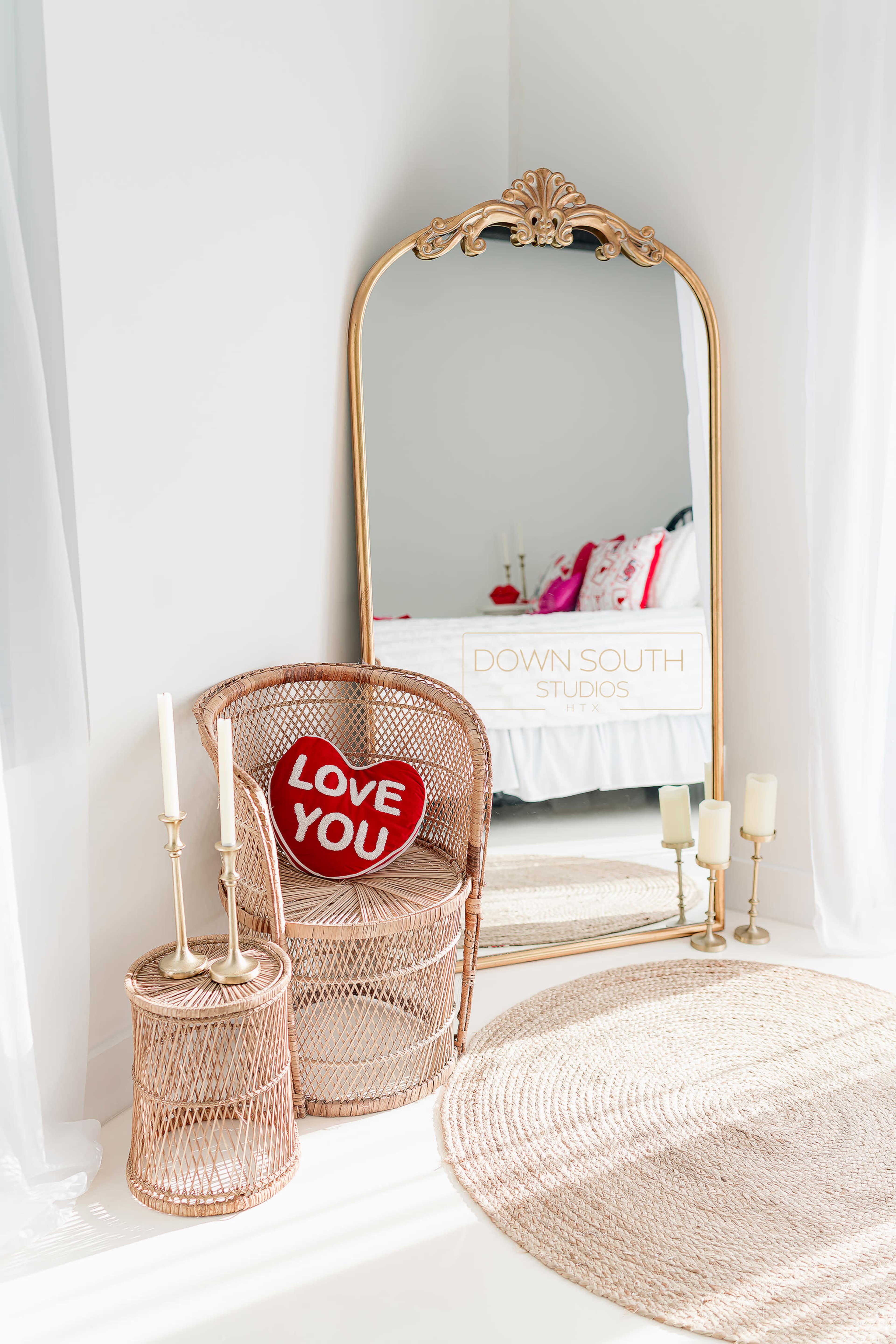 A large ornate mirror is positioned next to a wicker chair with a red heart-shaped cushion, surrounded by candle holders and a woven rug.