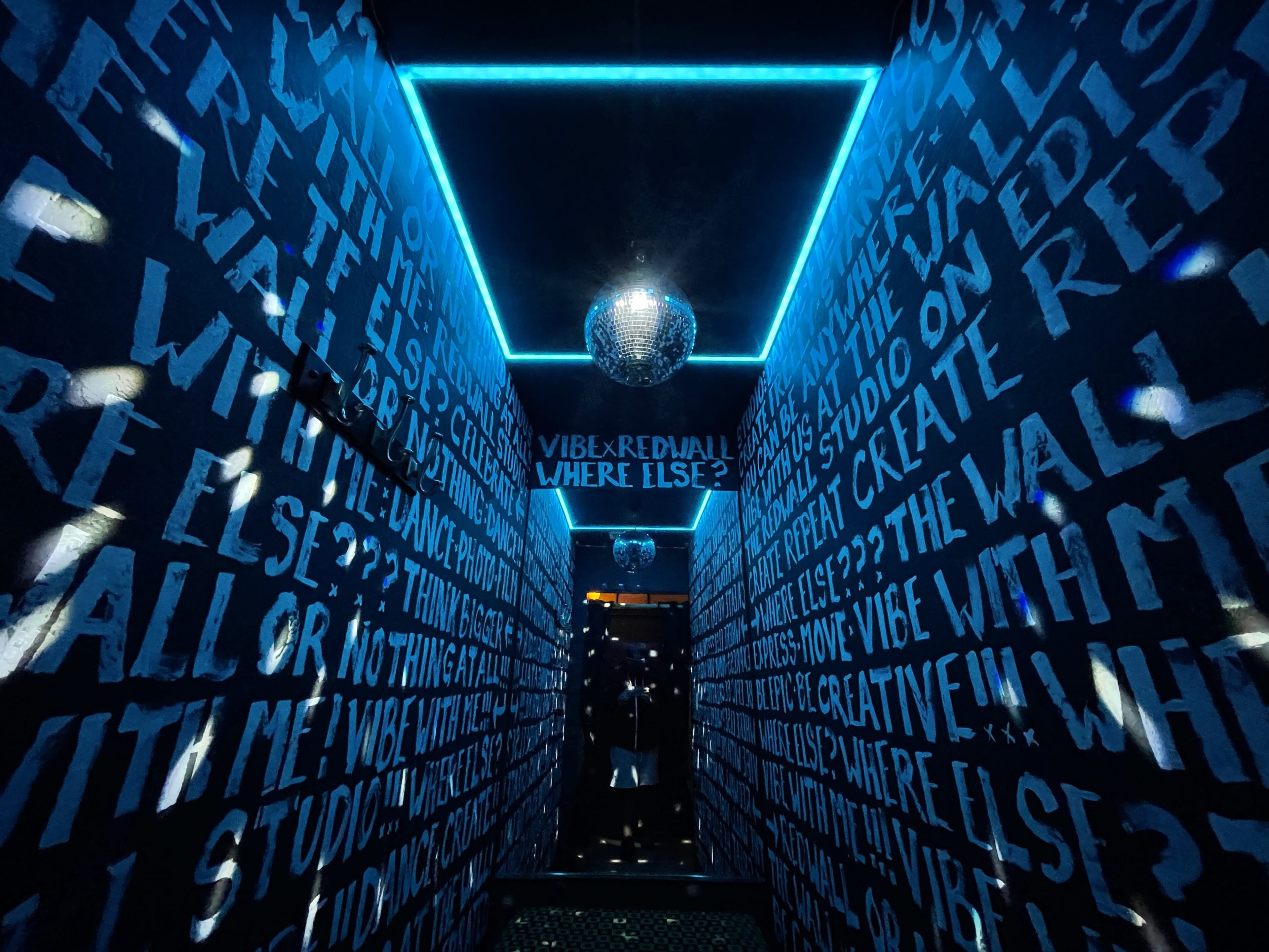A narrow hallway with walls covered in black paint featuring repeated white text, and a disco ball hanging from the ceiling reflecting light.