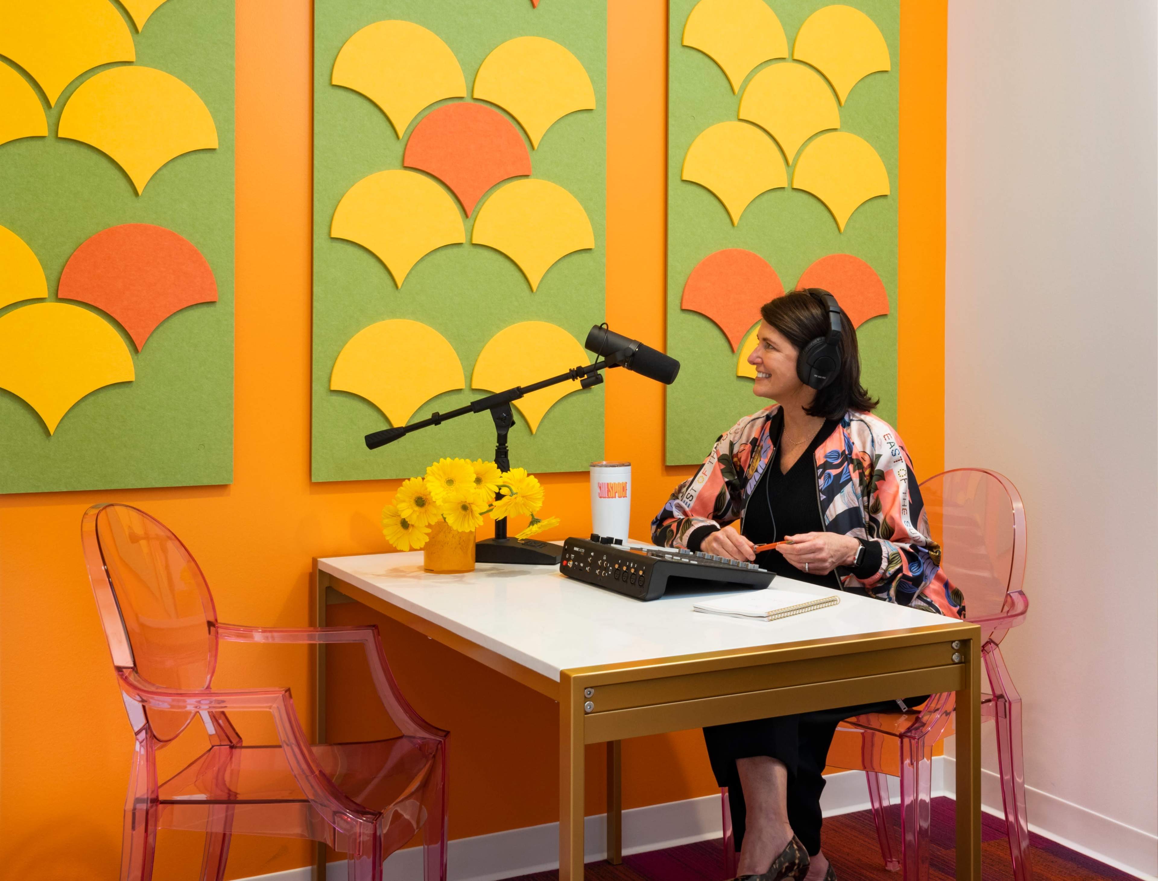 A woman sits at a small white desk with a microphone and headphones in a brightly colored recording studio featuring vibrant wall decor.