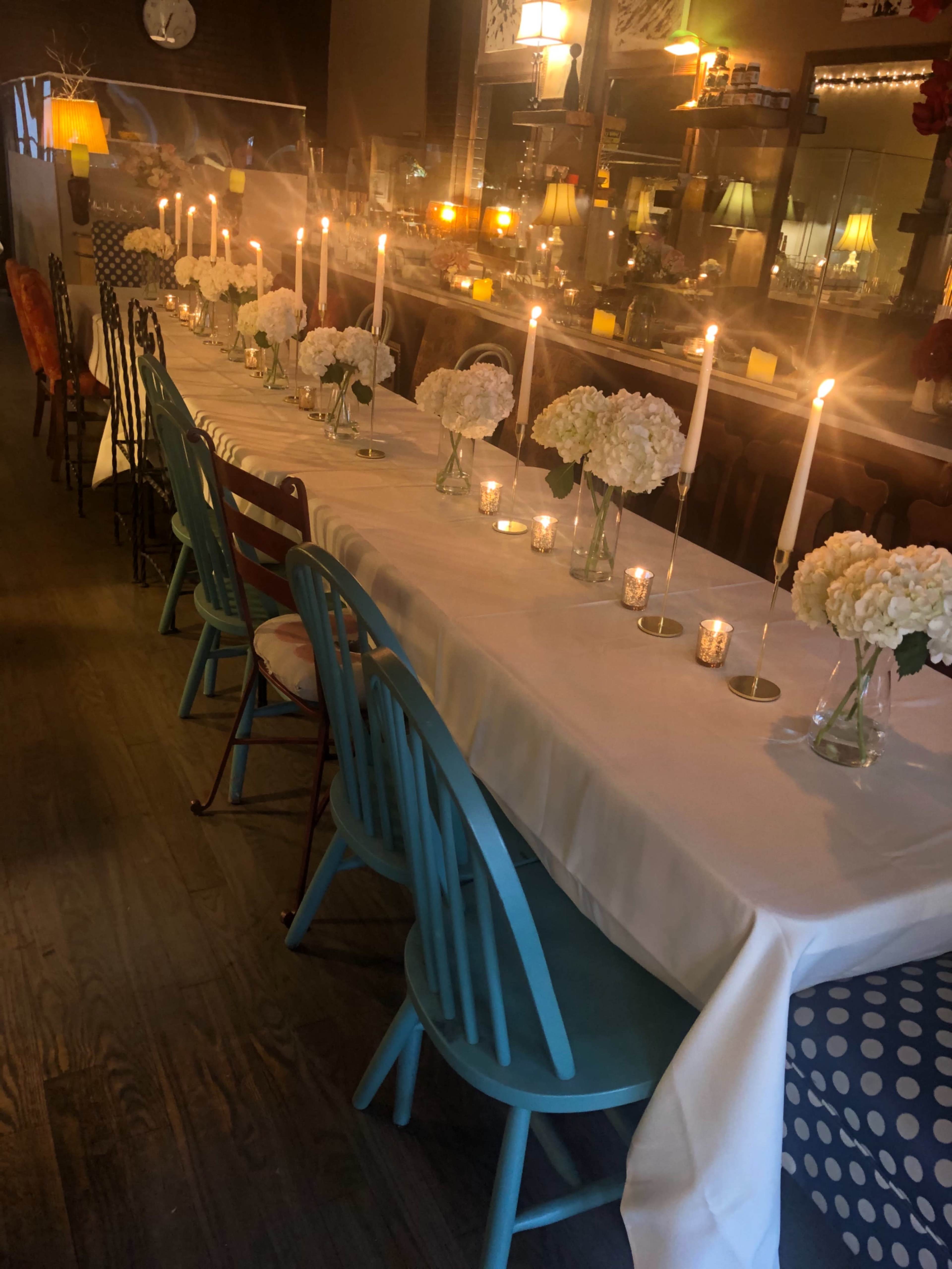 A long dining table is set with white flowers in glass vases and lit candles, surrounded by colorful chairs in a warmly lit restaurant.