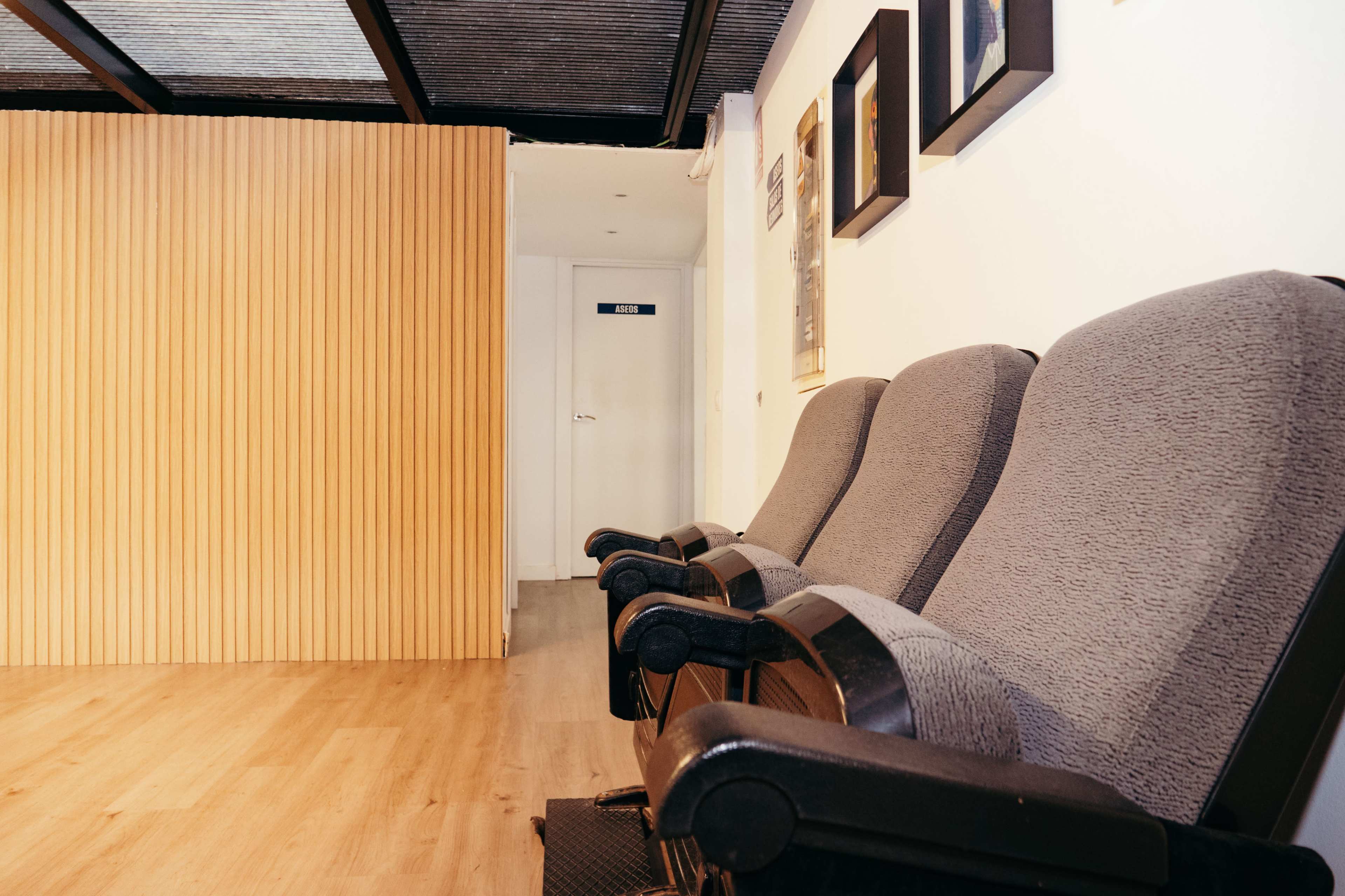 The image shows a waiting area with three gray chairs positioned against a wall, and a wooden partition leading to a door at the back.