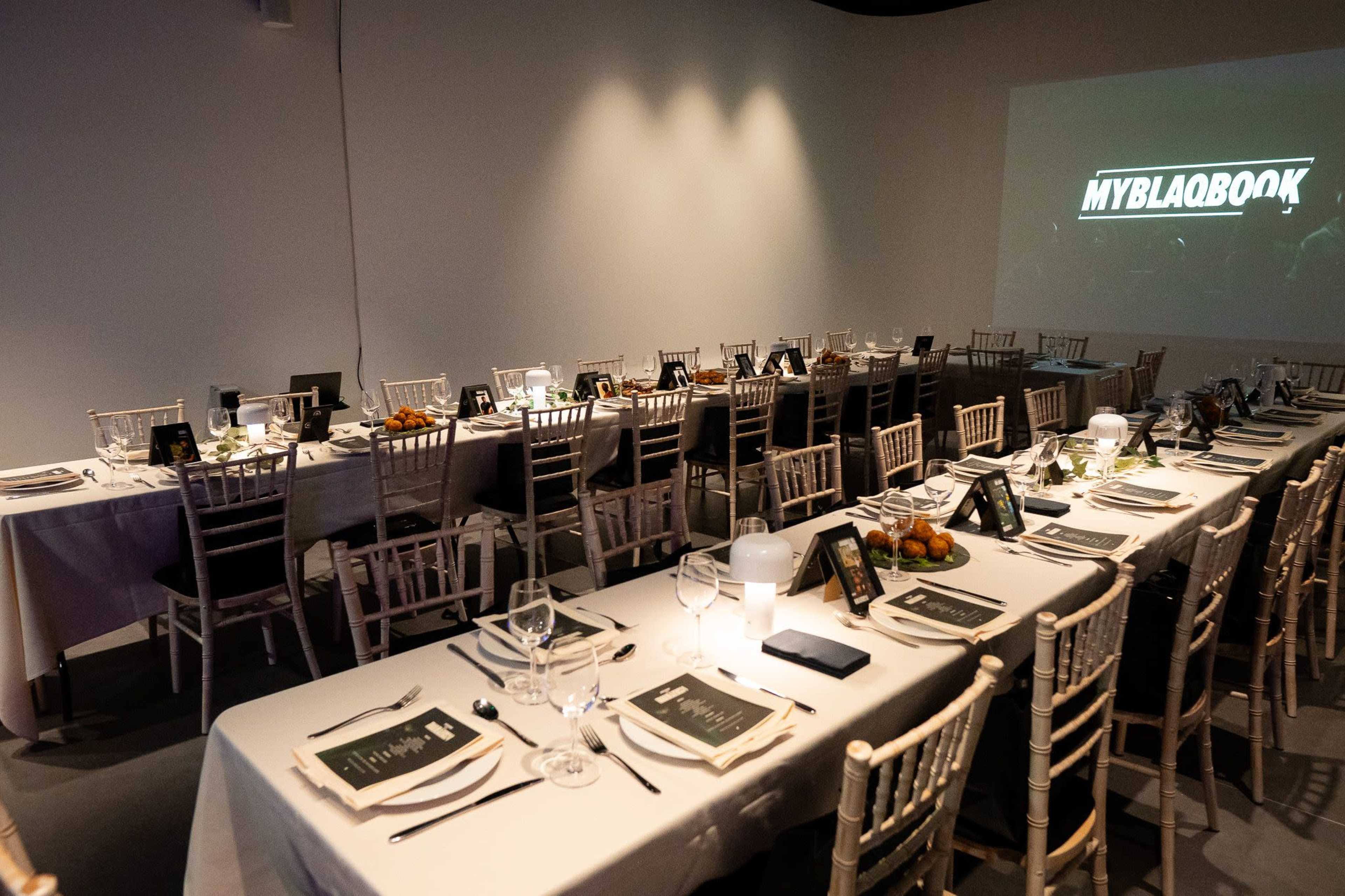 A long banquet table is set with elegant tableware and decorations, ready for an event, with a projector screen displaying "MYBLAQBOOK" in the background.