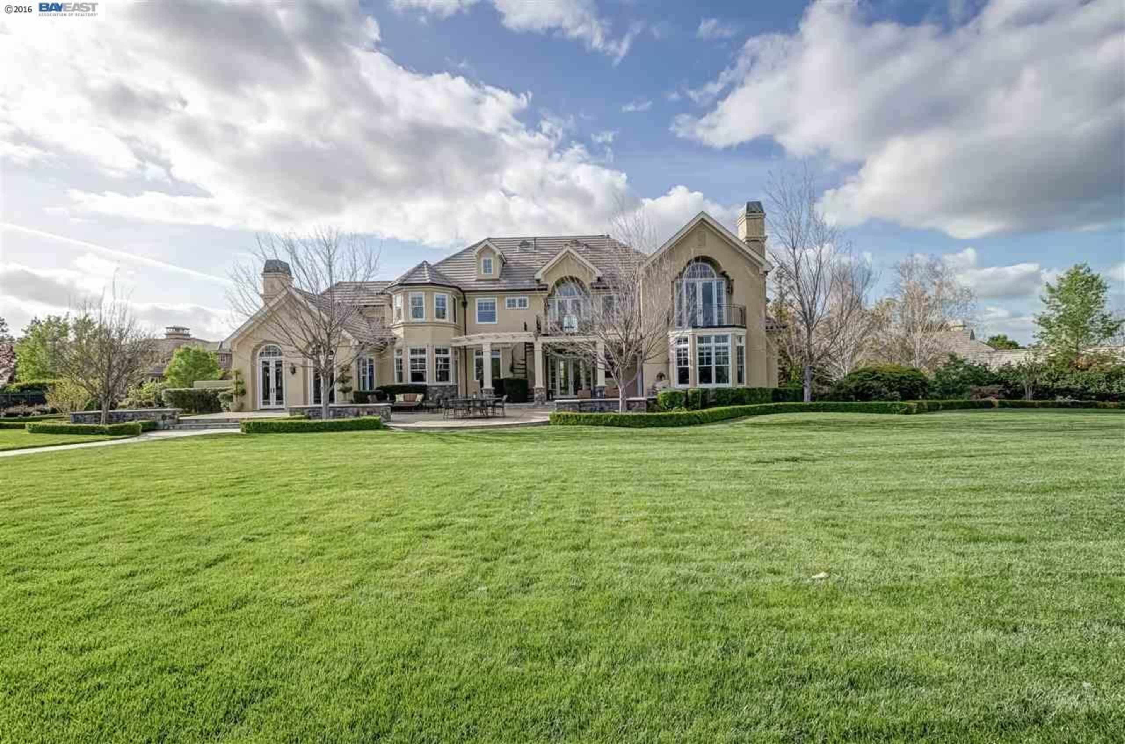 A large, two-story house with a manicured lawn and several trees sits under a partly cloudy sky.