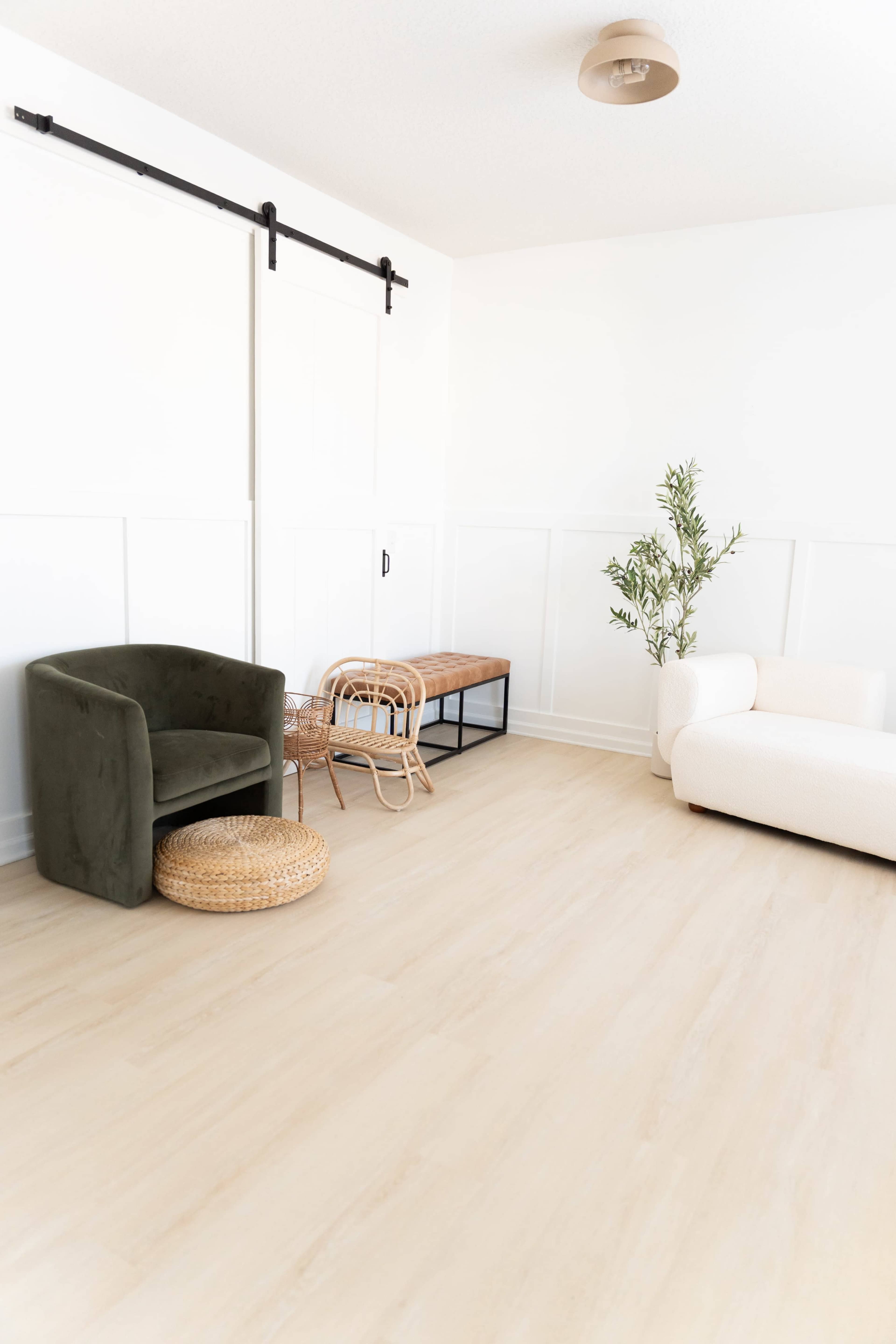 The image shows a minimalist room with light wood flooring, featuring a green upholstered chair, a woven stool, a wooden bench, and a potted plant.