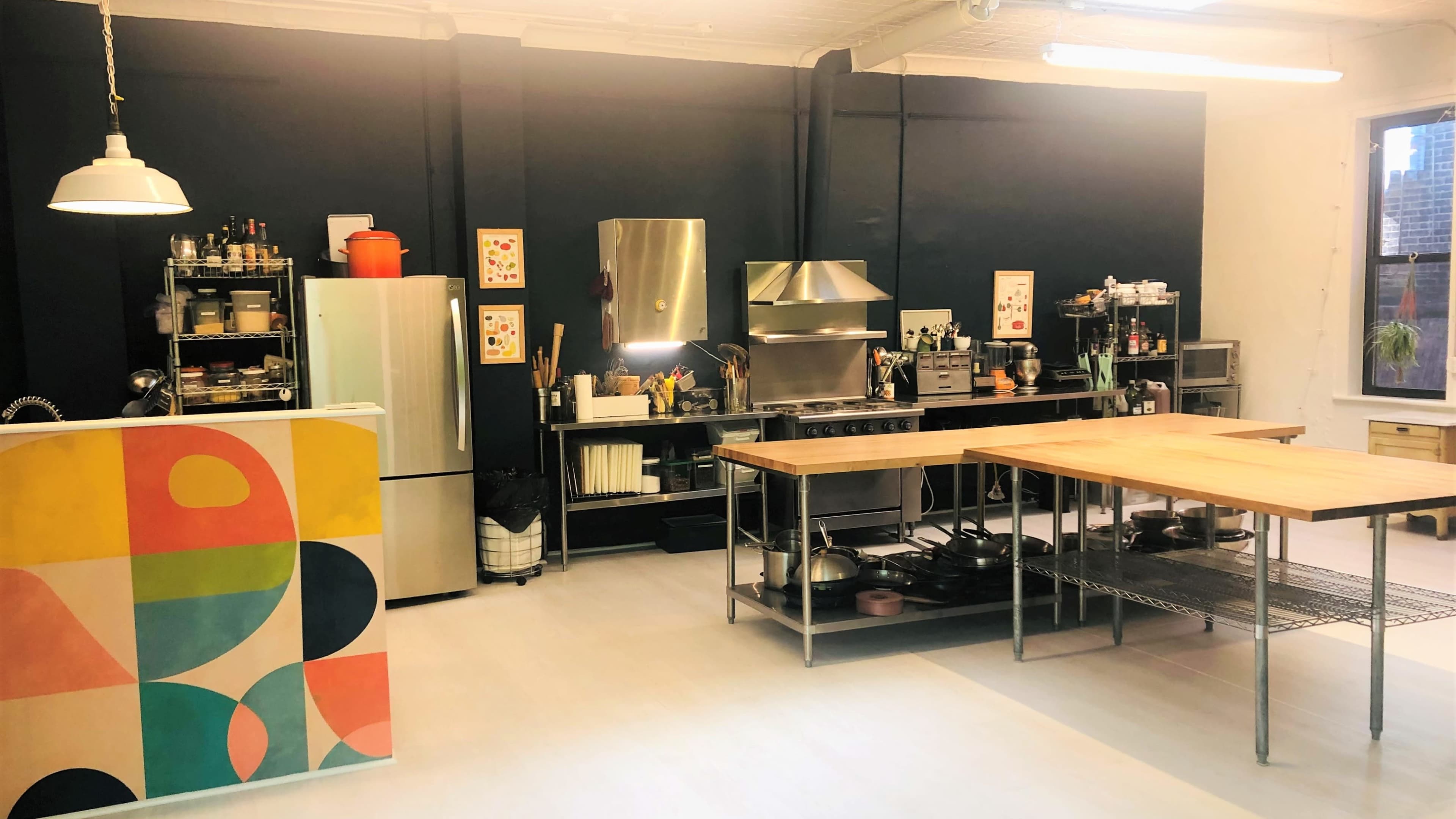 A modern kitchen with a stainless steel stove, a refrigerator, and multiple work surfaces arranged in an organized manner against a dark blue wall.