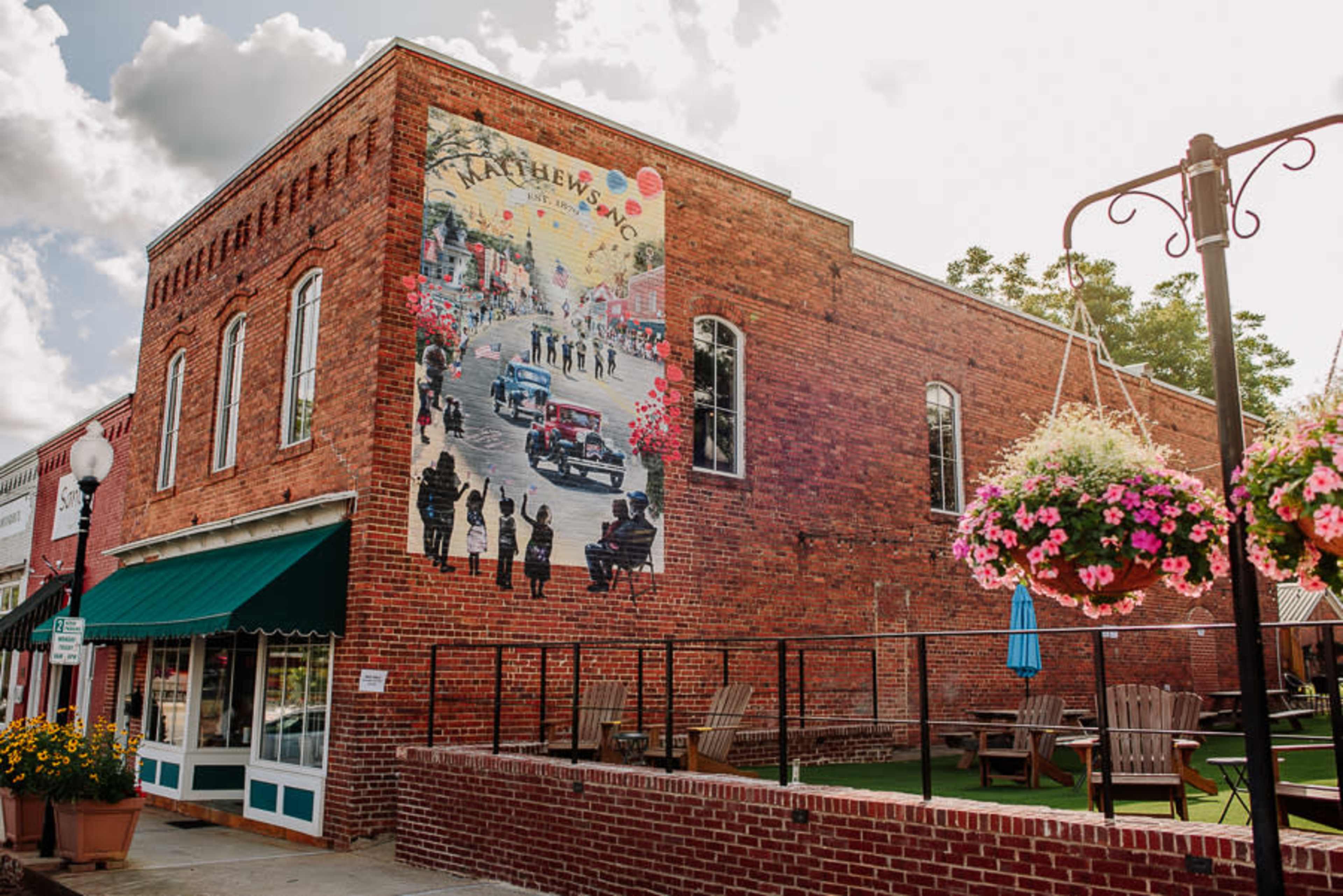 A brick building features a colorful mural depicting a historic street scene, with flower planters and outdoor seating visible in front.