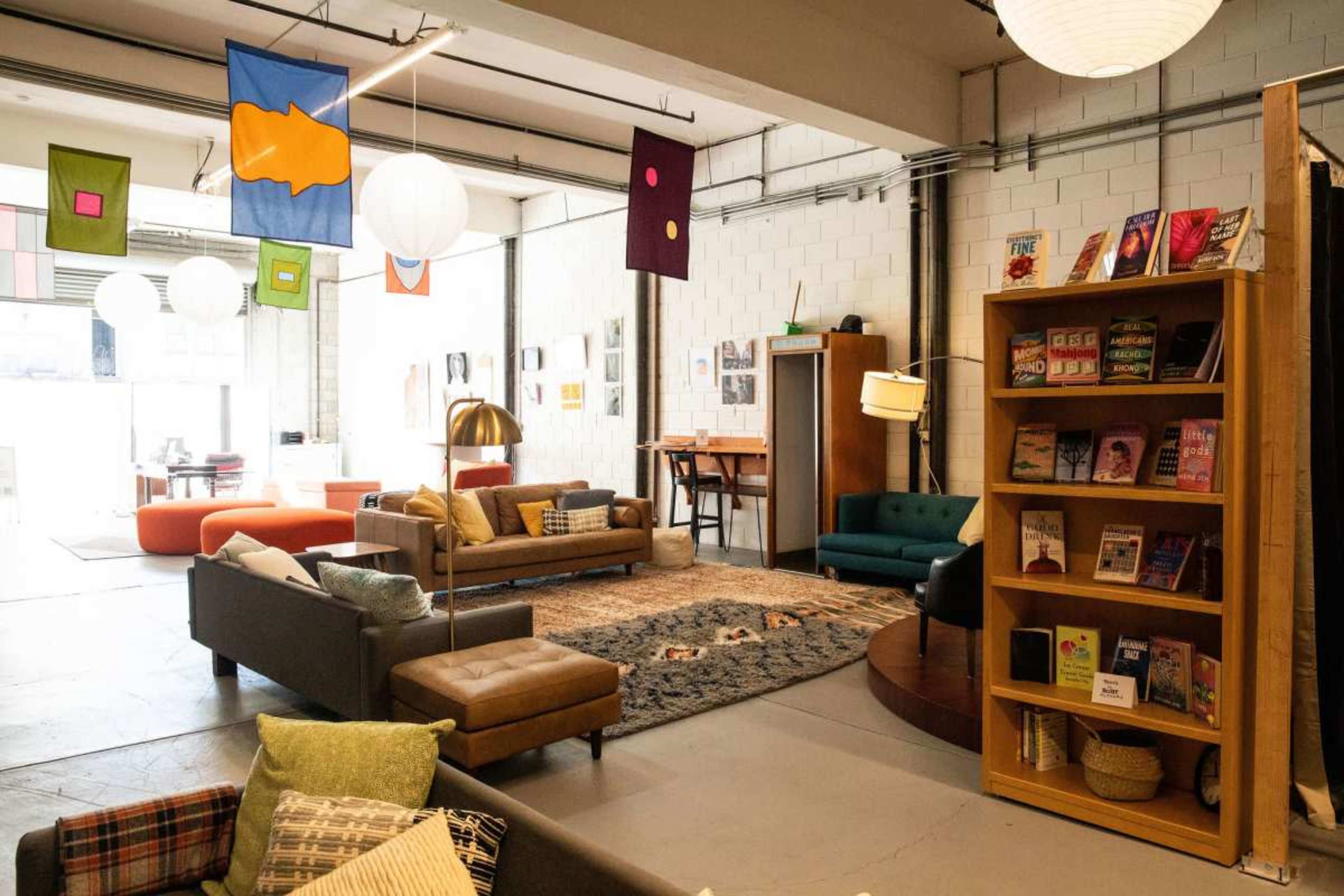 The image shows a modern, open lounge area featuring various seating options, colorful wall hangings, and bookshelves in a well-lit space.