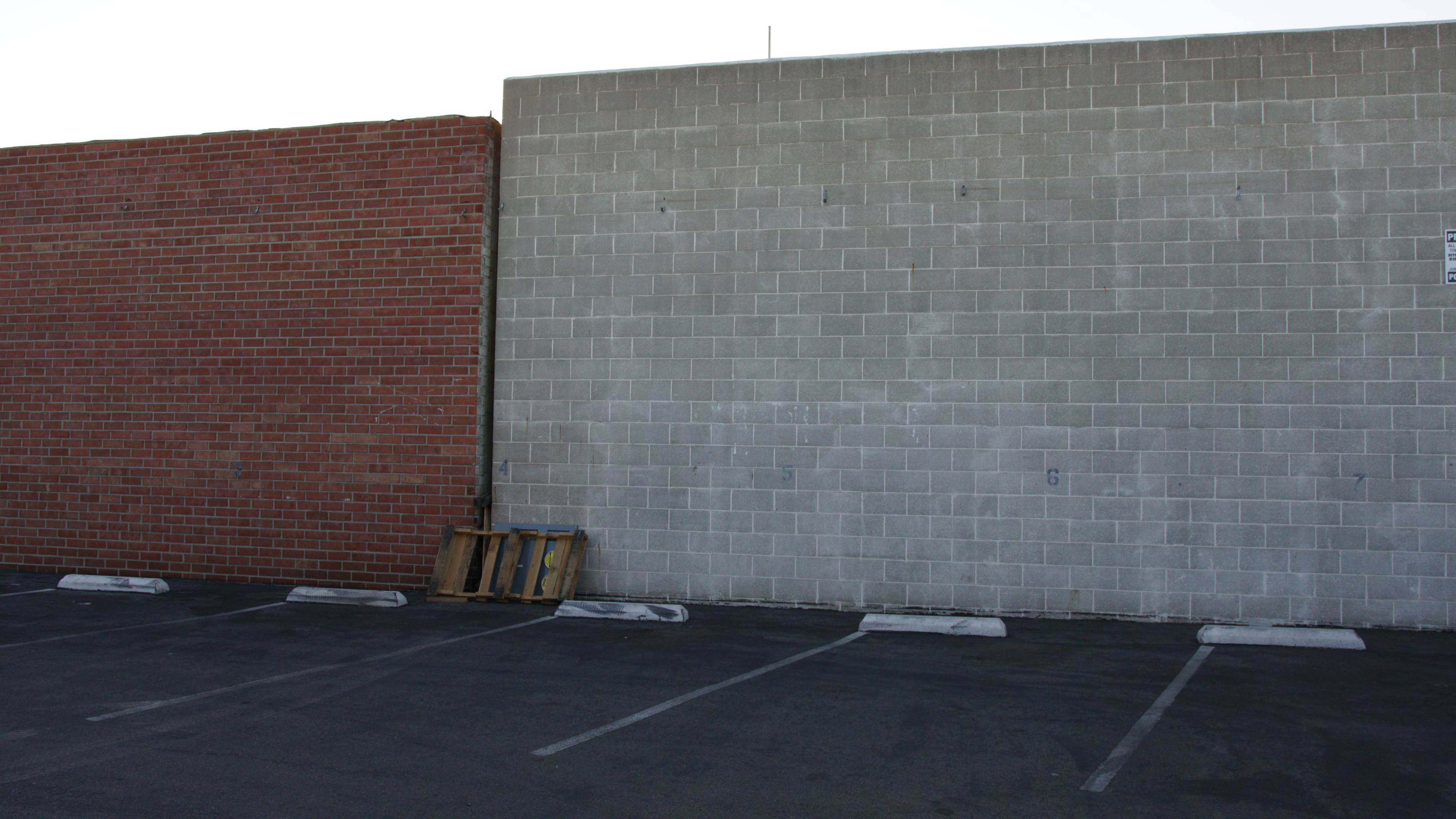 A parking lot with a gray concrete wall on the right and a red brick wall on the left, with a wooden pallet leaning against the gray wall.