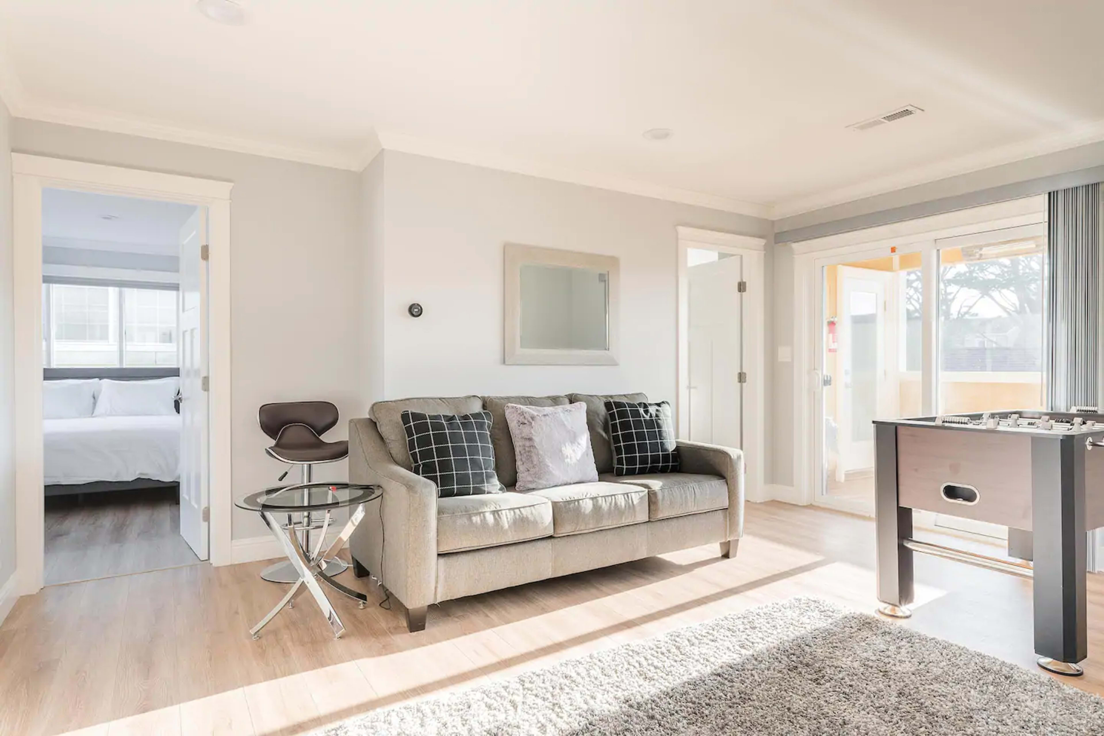 A well-lit living room features a gray sofa with decorative pillows, a small glass table, and a foosball table, with a doorway leading to a bedroom and sliding glass doors that open to a balcony.