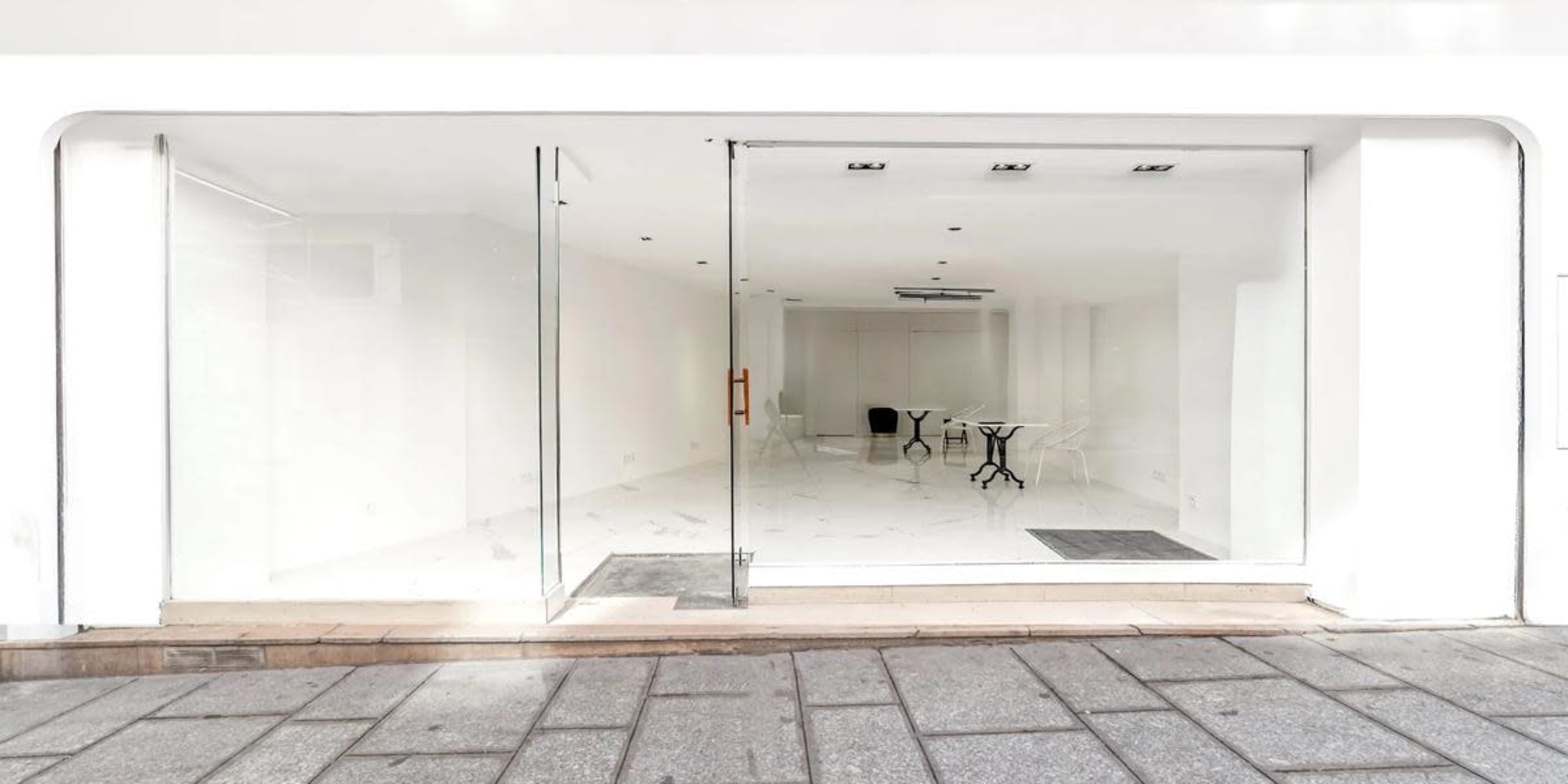The image shows a minimalist glass storefront with an empty, well-lit interior featuring a small table and chair.